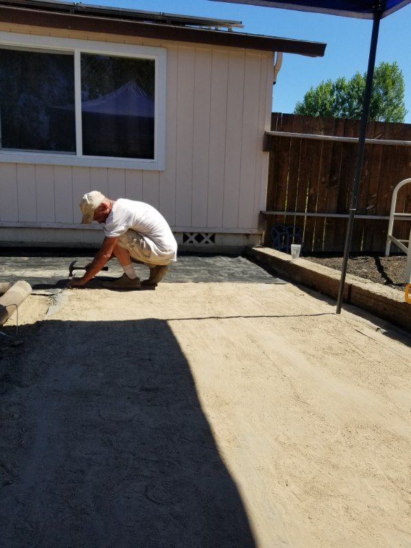 Worker kneeling on a concrete patio, smoothing wet cement beside a house in bright sunlight