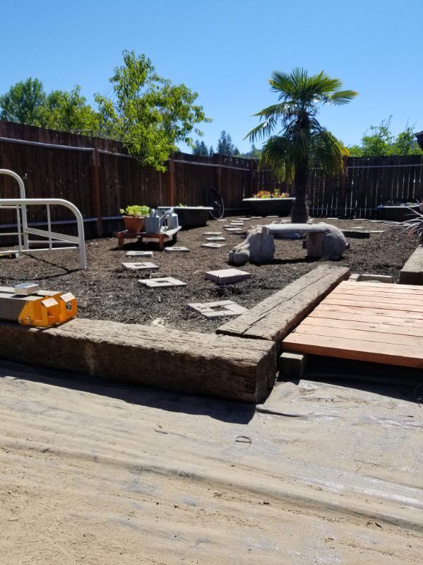 Backyard under construction with gravel, pavers, a wooden deck, and a fence in bright sunlight
