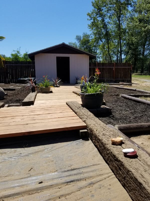 Backyard with a wooden deck walkway leading to a small white shed, raised garden beds, and trees beyond.