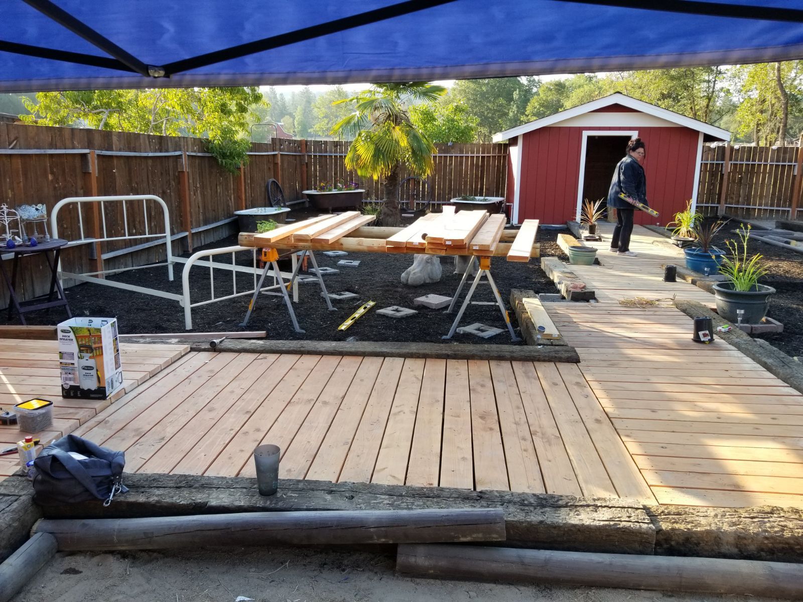 Backyard deck under construction with red shed, lumber stacks, and a person working near the fence