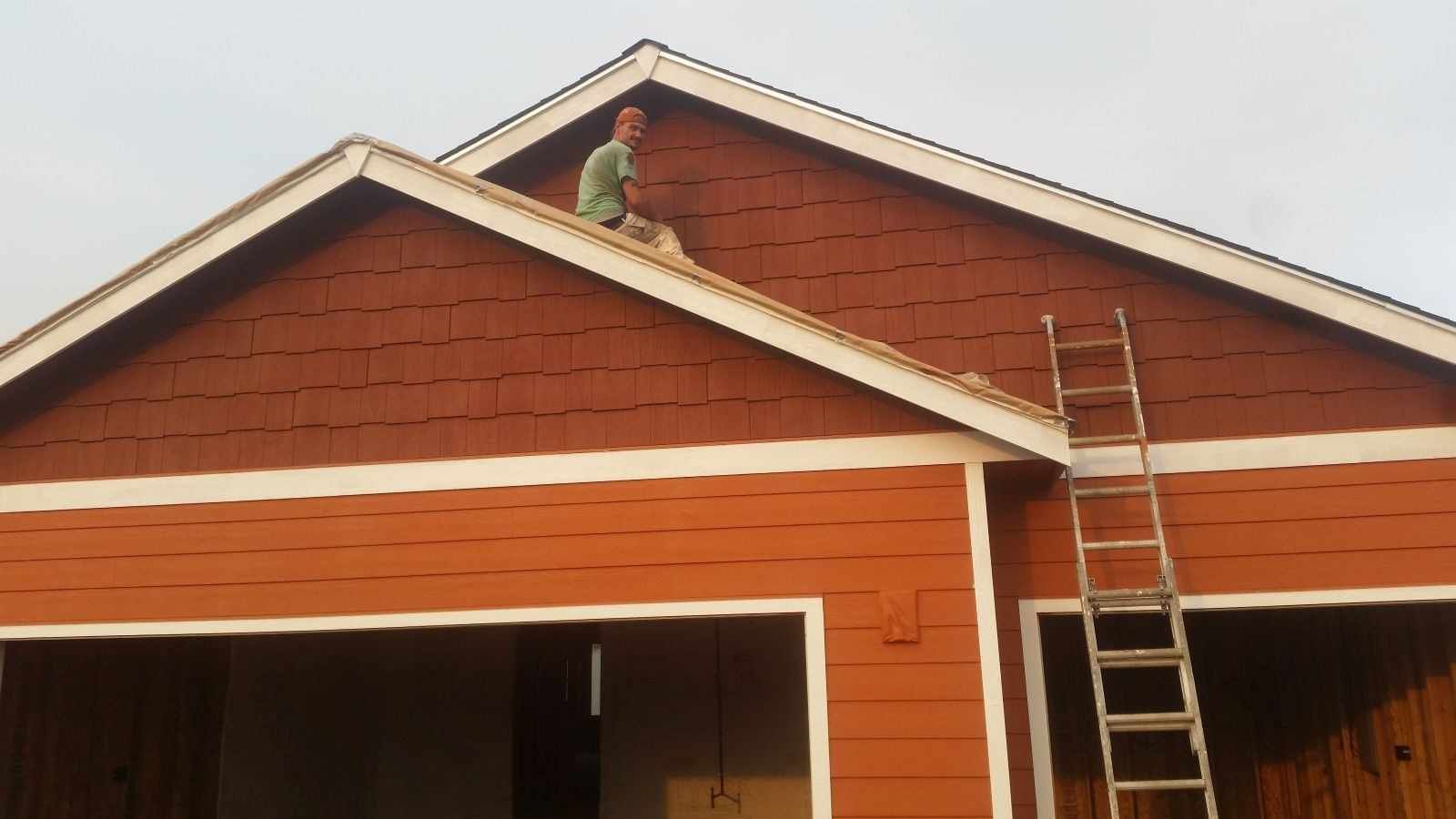 Worker on a roof beside a ladder, painting a red house gable with white trim.