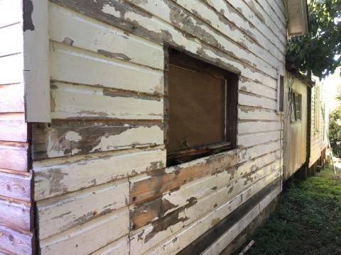 Peeling white house siding with a boarded-up window and weathered paint