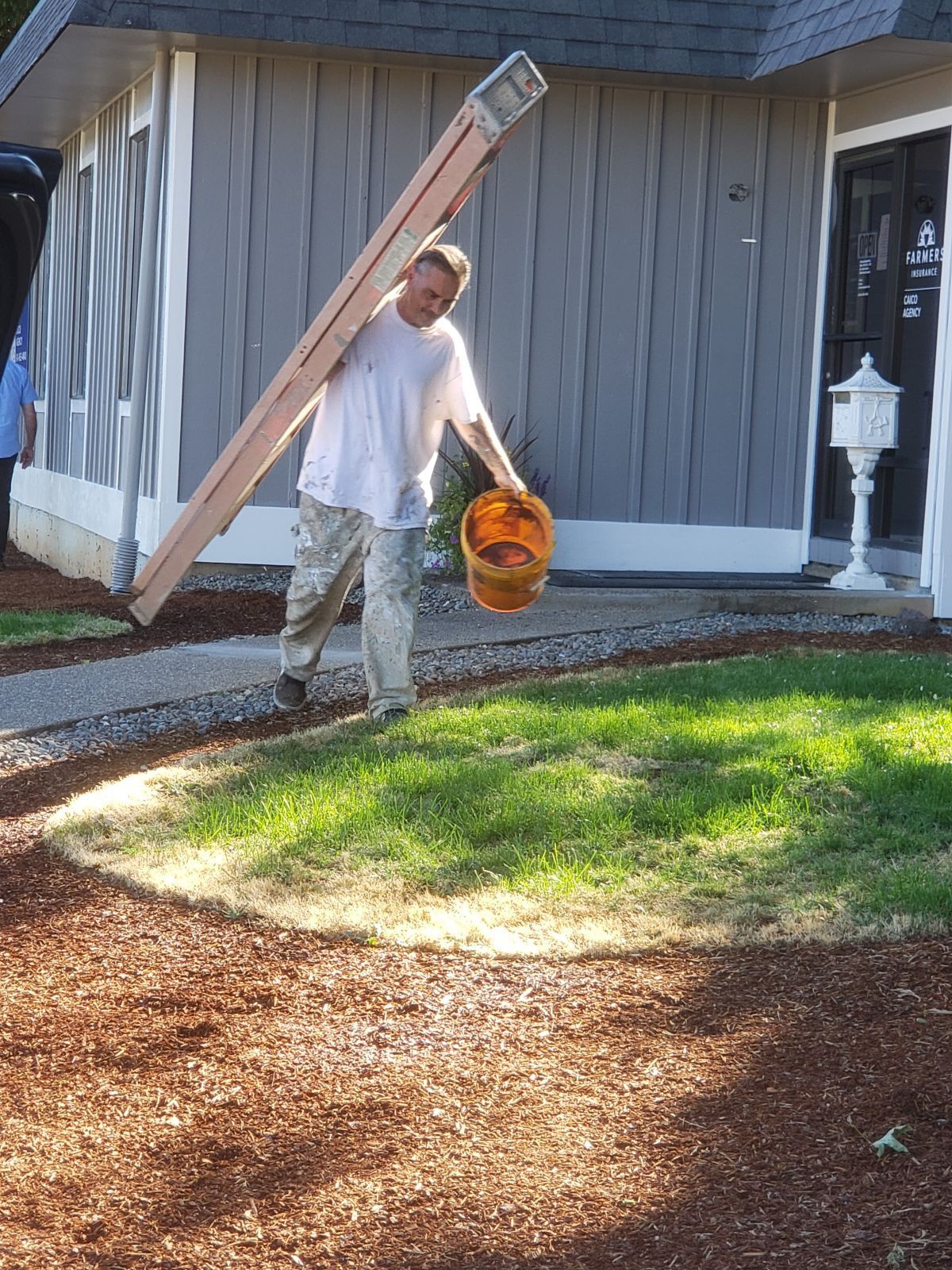 Man carrying a long board and orange bucket near a house, standing on a grassy yard.
