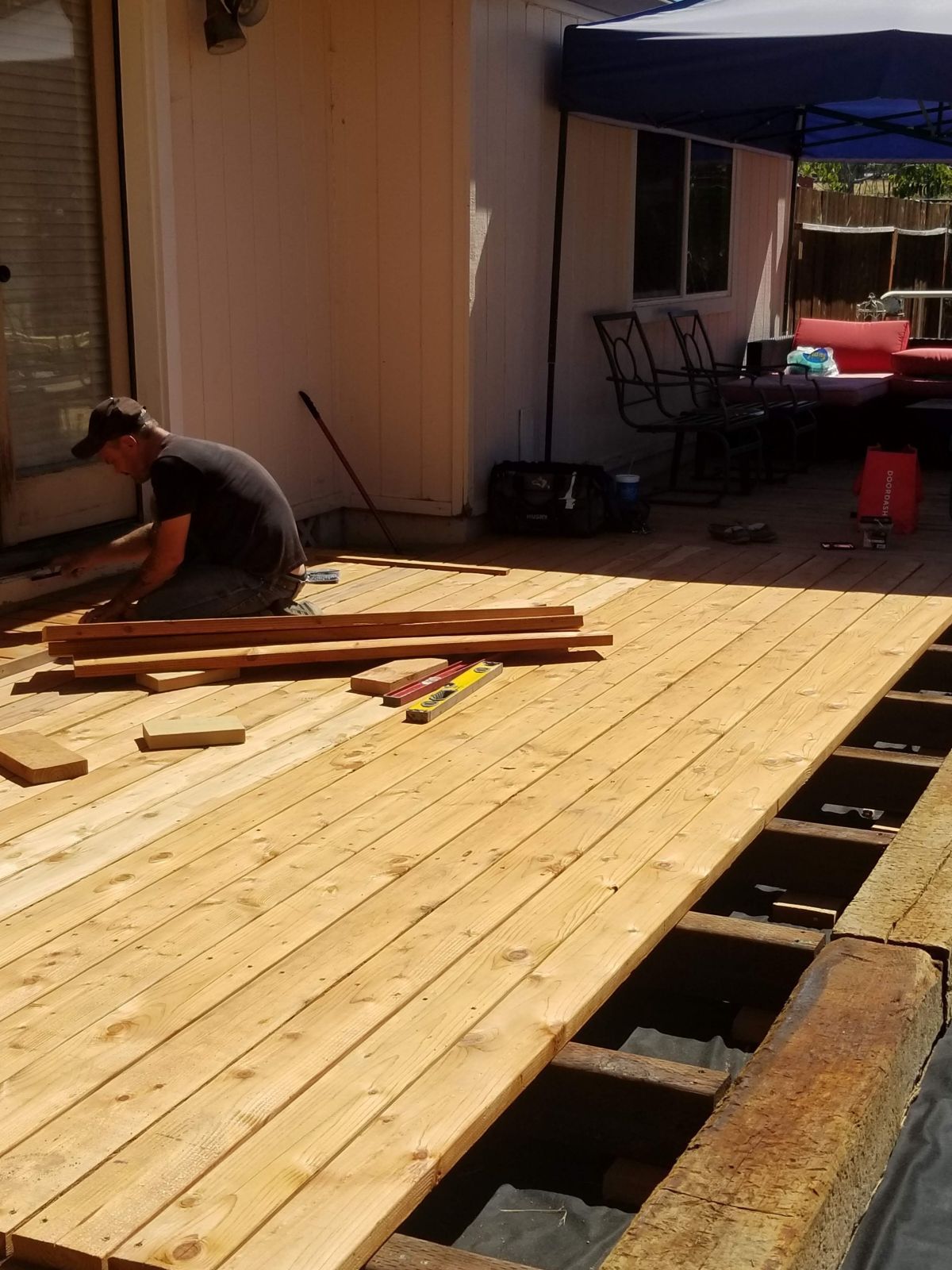Worker building a wooden deck beside a house, with exposed joists and tools on the boards.