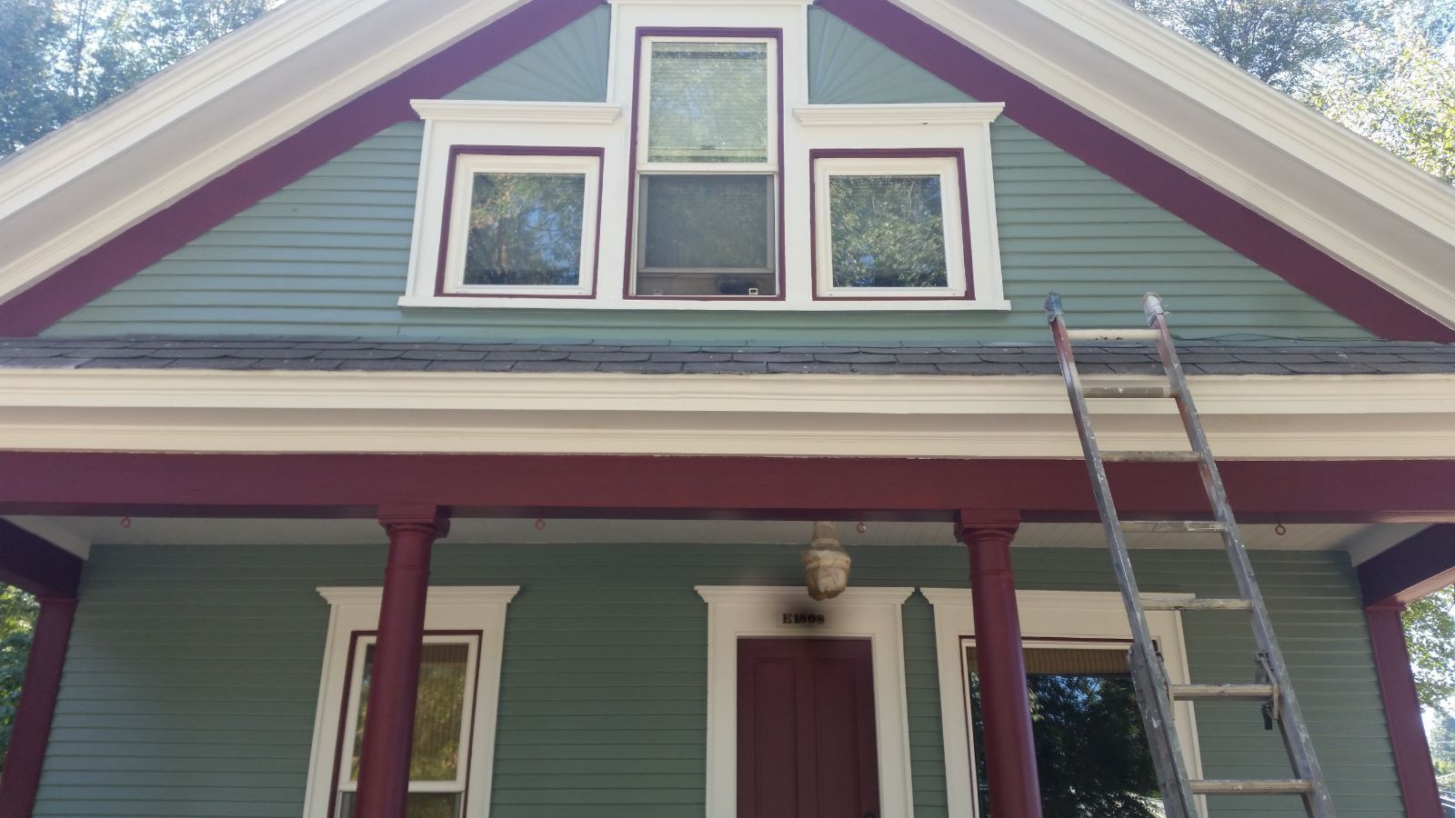 Front of a house with green siding, red trim, three windows, and a ladder leaning against the porch roof.