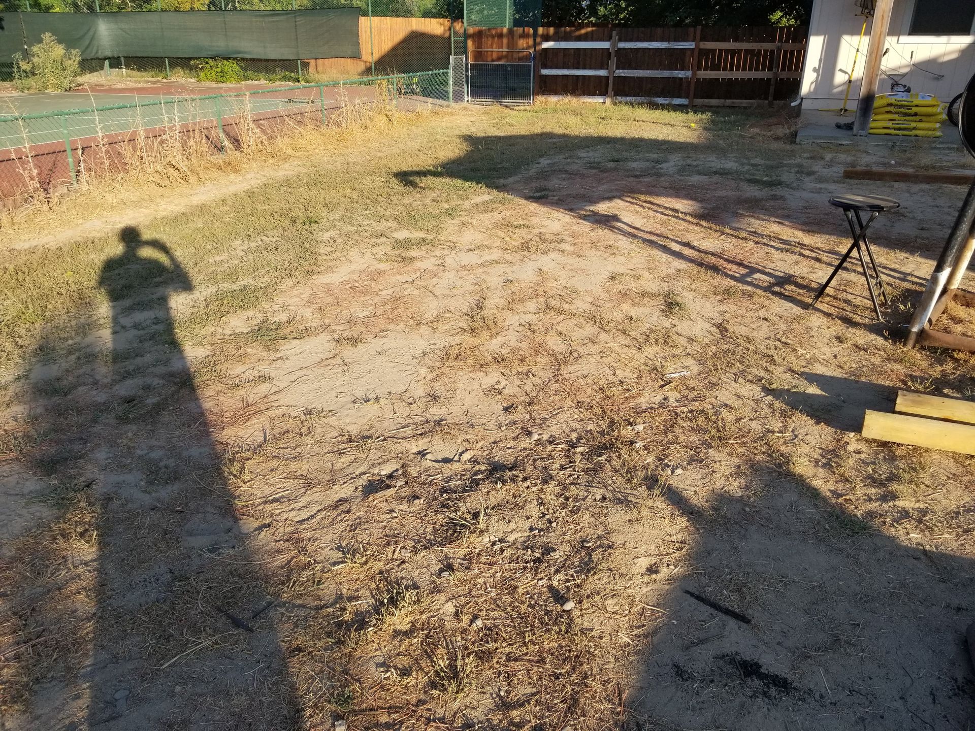 Sunlit backyard with patchy dirt and grass, a fence, and a patio table casting long shadows