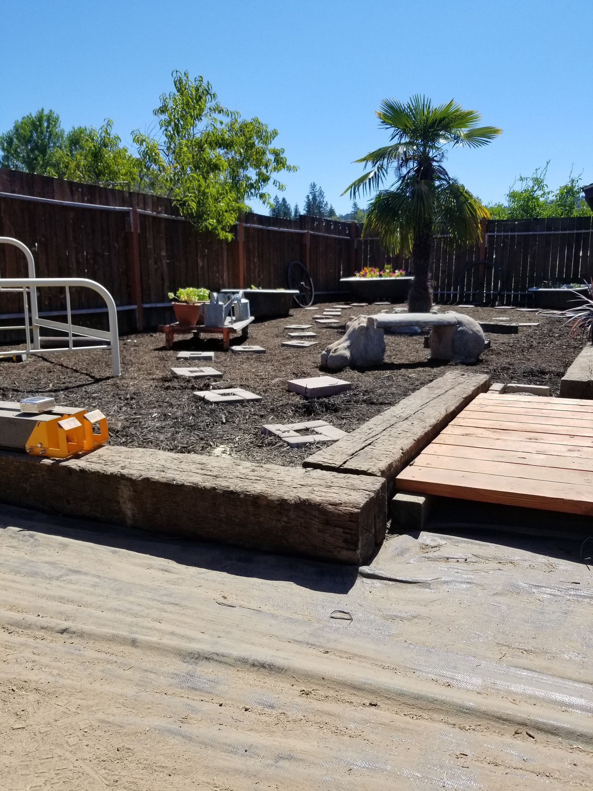 Backyard with raised gravel landscaping, boulders, a wooden deck, and scattered tools beside a fence