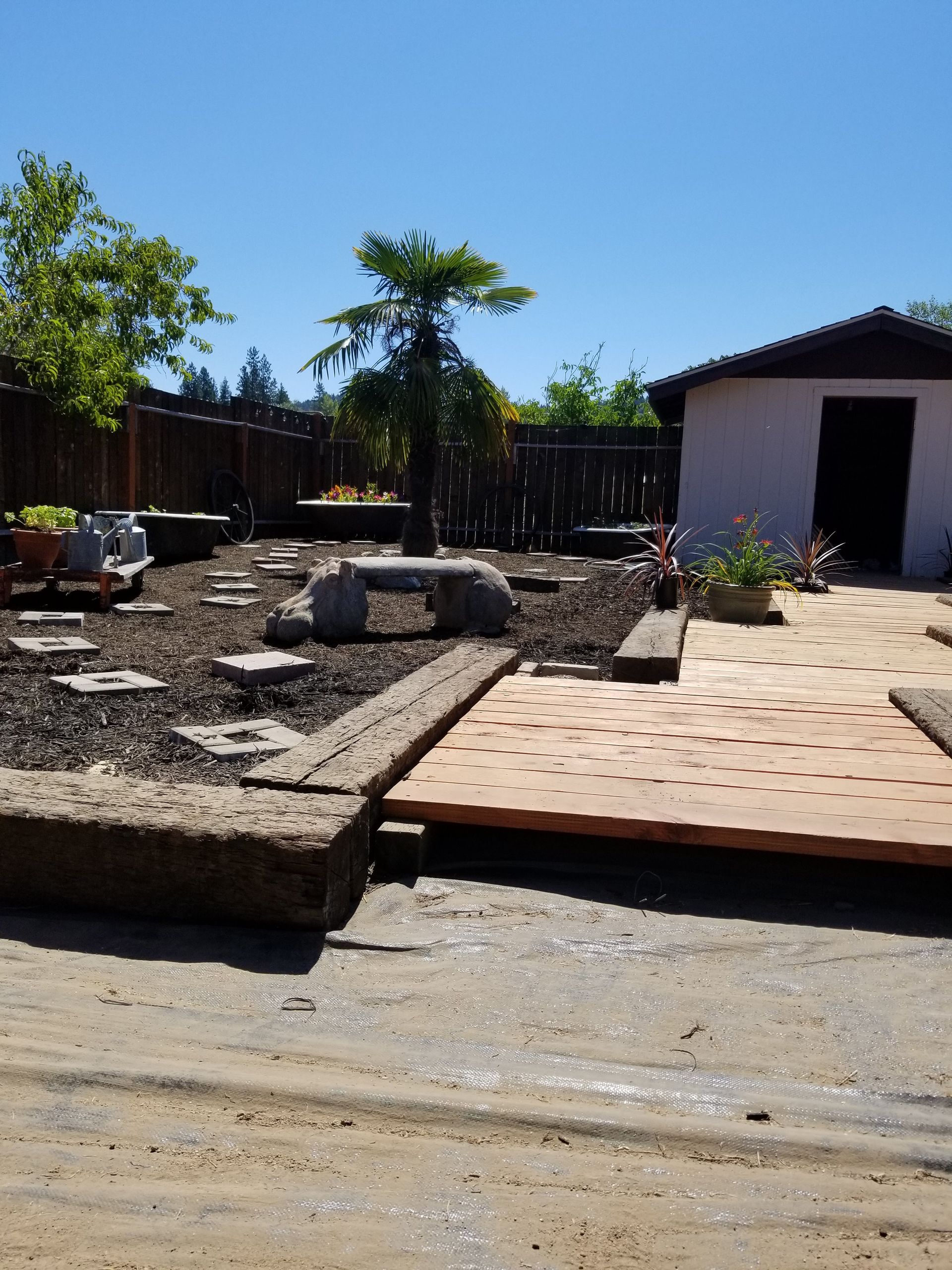 Sunlit backyard with stone patio, gravel beds, and a small white shed under a clear blue sky
