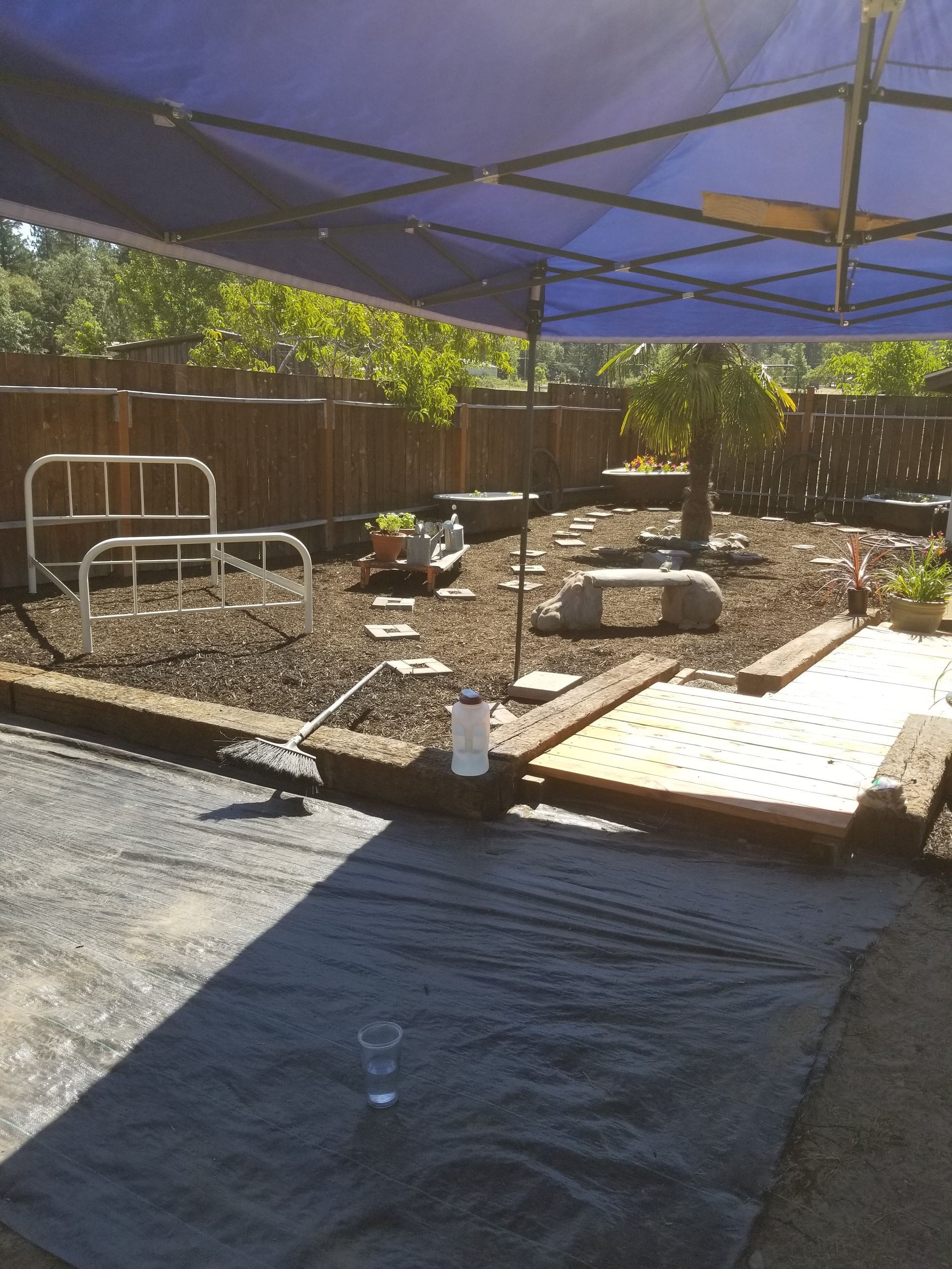 Shaded backyard construction area with dirt, gravel, lumber, and a blue canopy overhead.