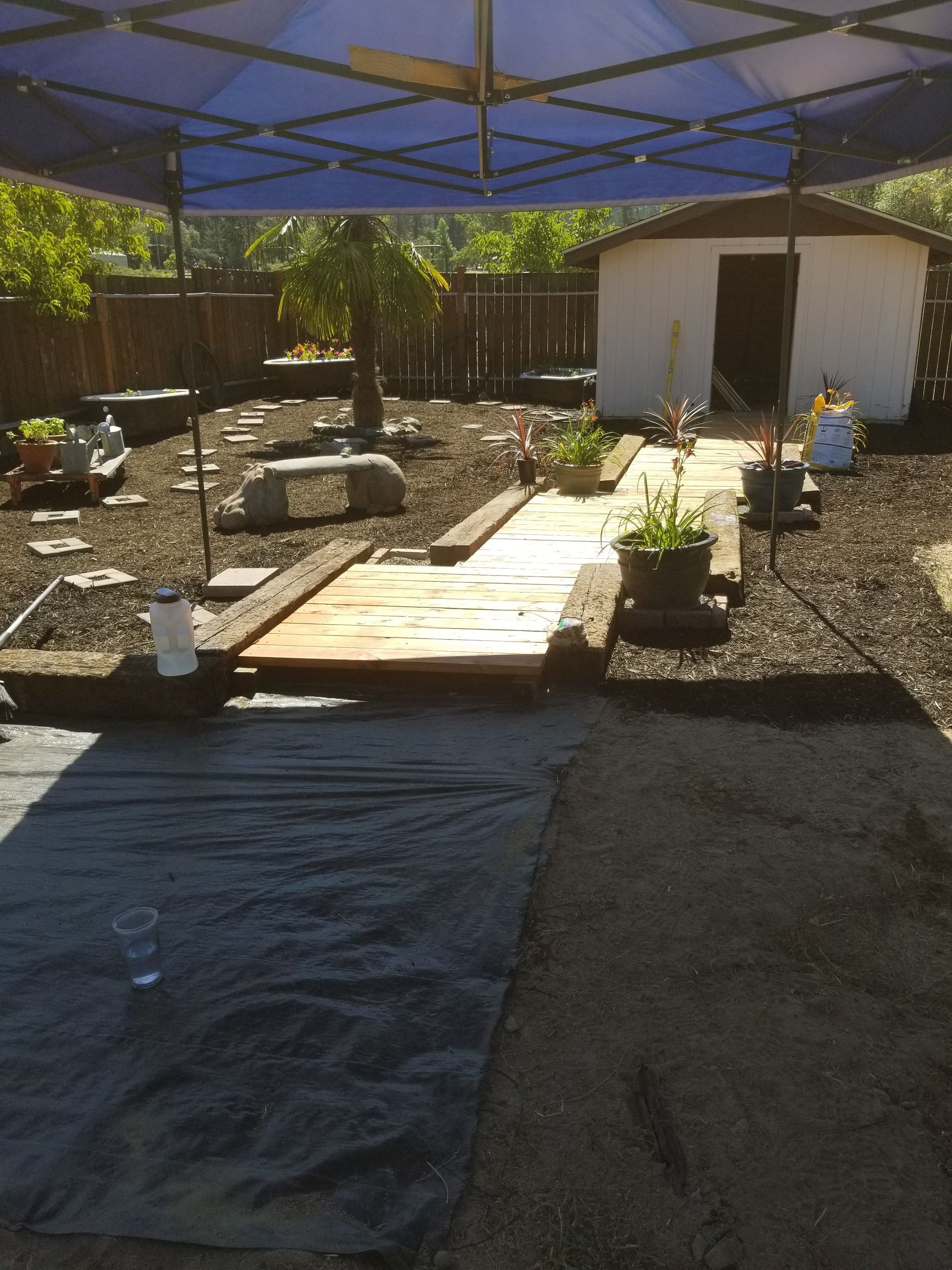 Sunlit backyard with canopy, gravel path, wooden platform, and a small shed or outbuilding.