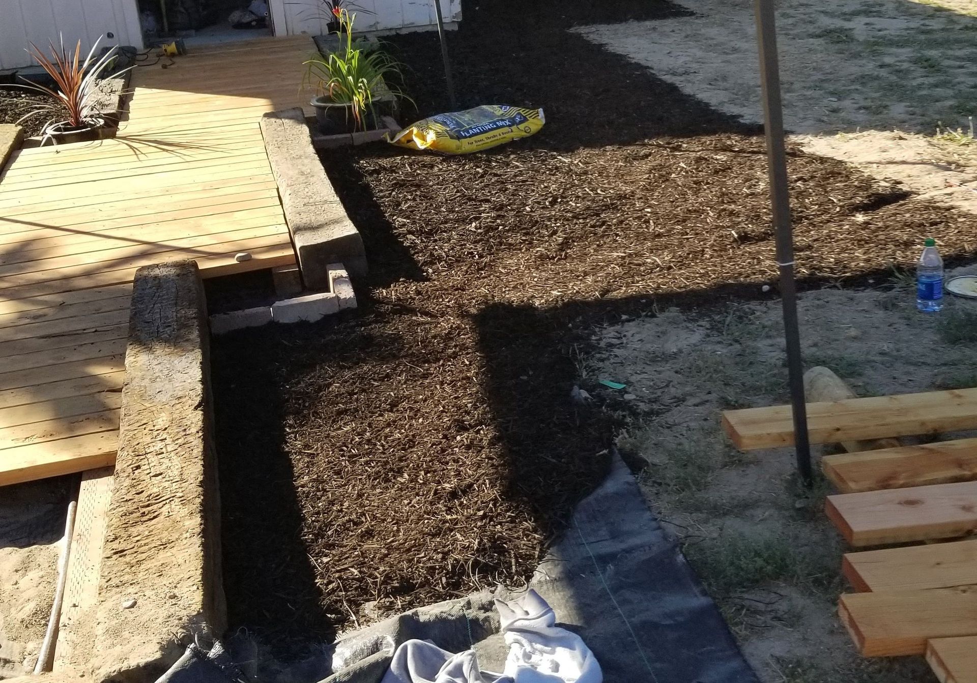Garden path under construction beside a wooden deck, with gravel, black fabric, and pavers laid out
