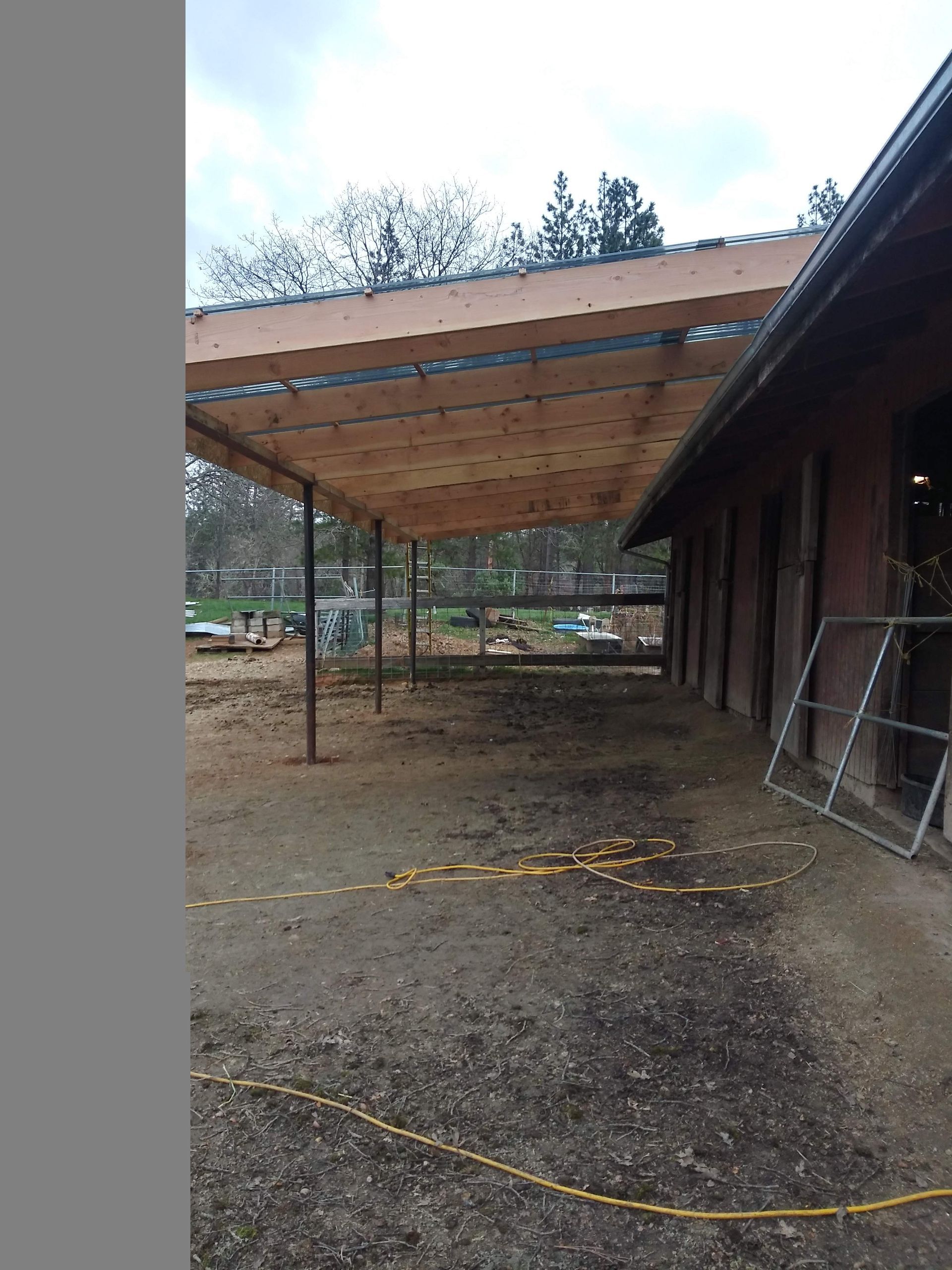 Wooden carport with metal posts beside a building on a muddy gravel lot, with trees in the background.