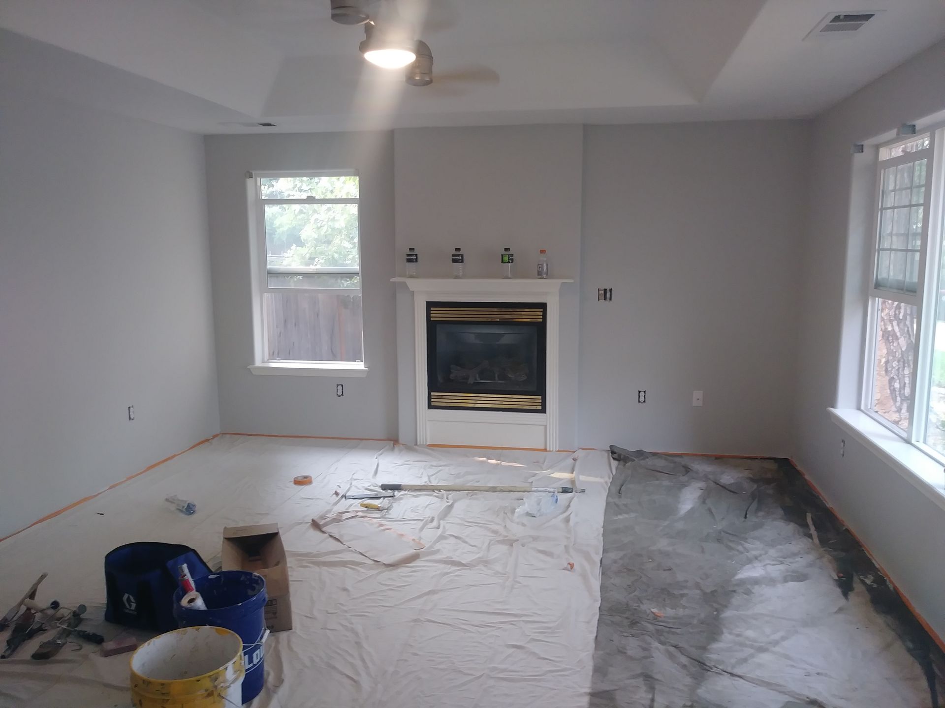 Empty room being renovated, with covered floors, a fireplace, and windows letting in daylight.