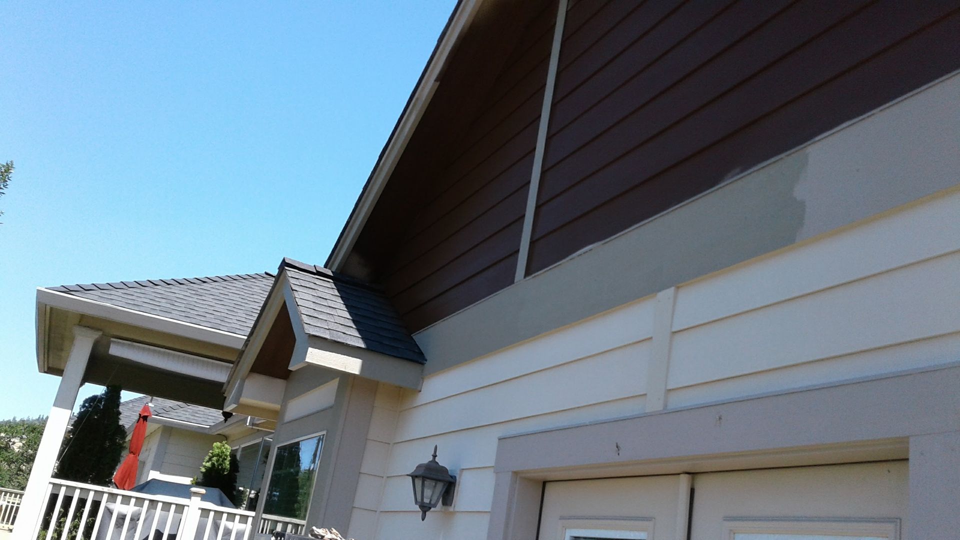 Suburban house exterior with beige siding, brown roof, and white fence under a blue sky