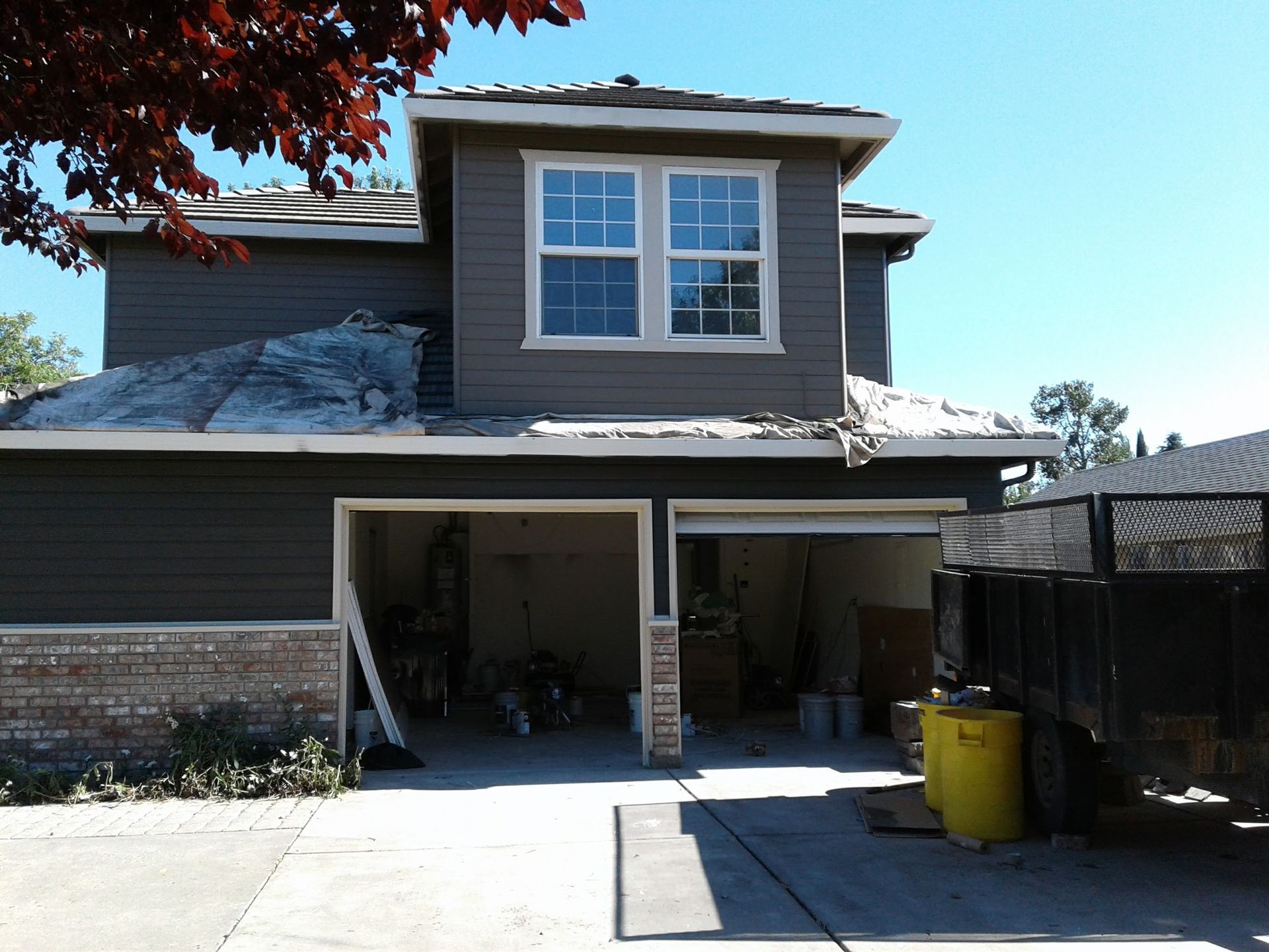 Two-story house with open garage, driveway, and yellow trash bin on a sunny day