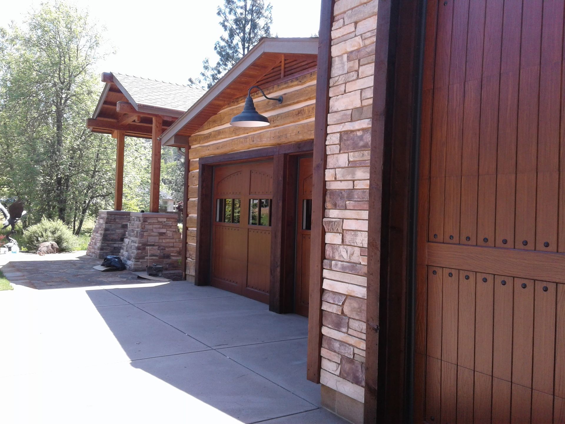 Wooden garage entrance with stone pillars and a shaded driveway beside a rustic house