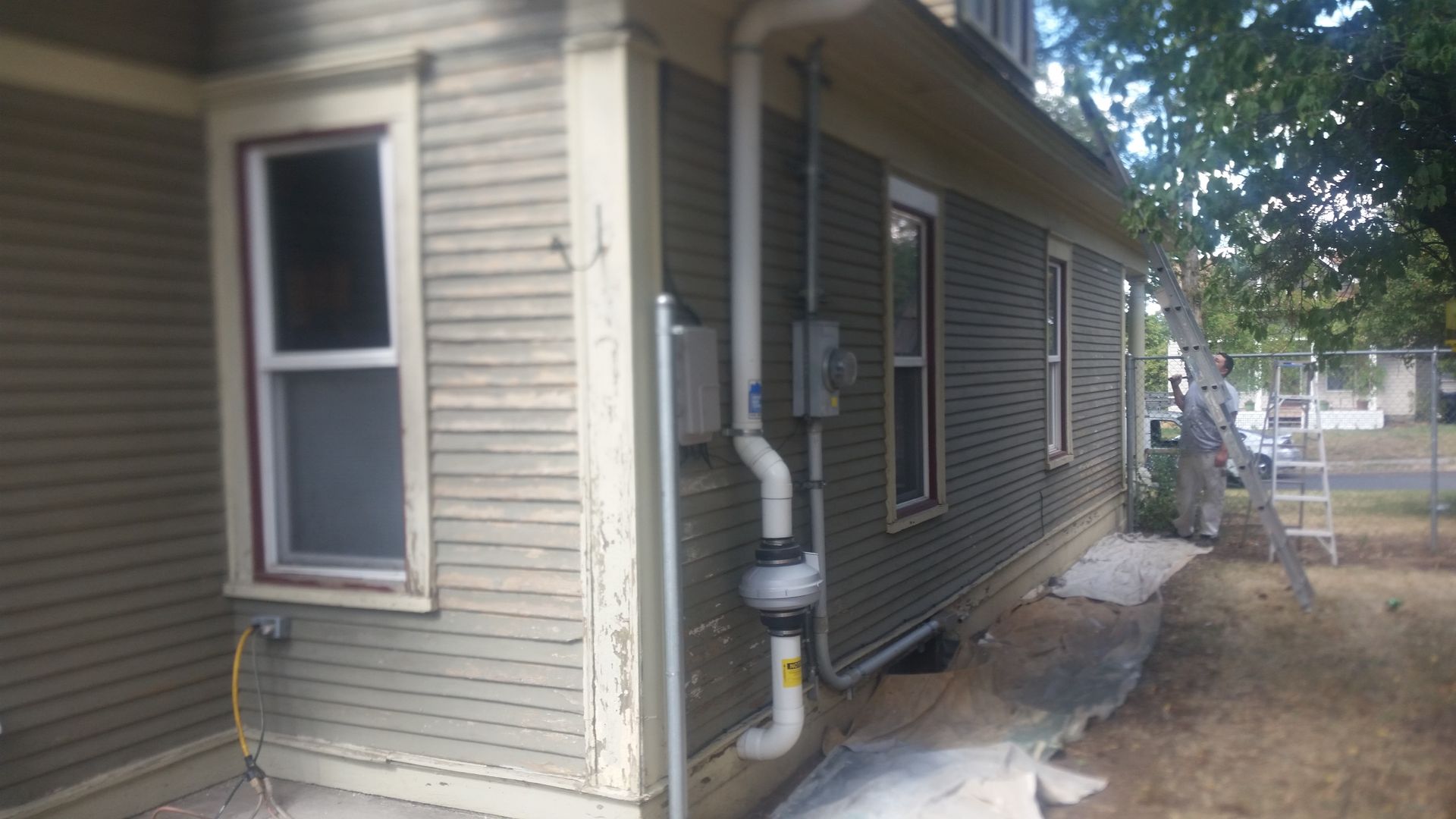 Side of a beige house with windows, utility meters, and a small concrete walkway beside a dirt yard.