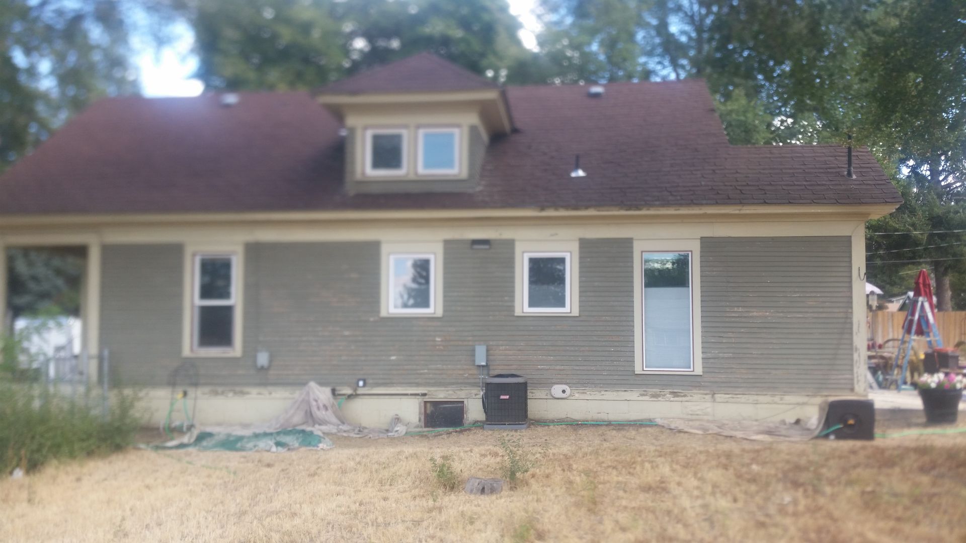 Single-story beige house with brown roof and dormer, front yard, and patio furniture in a dry grassy setting