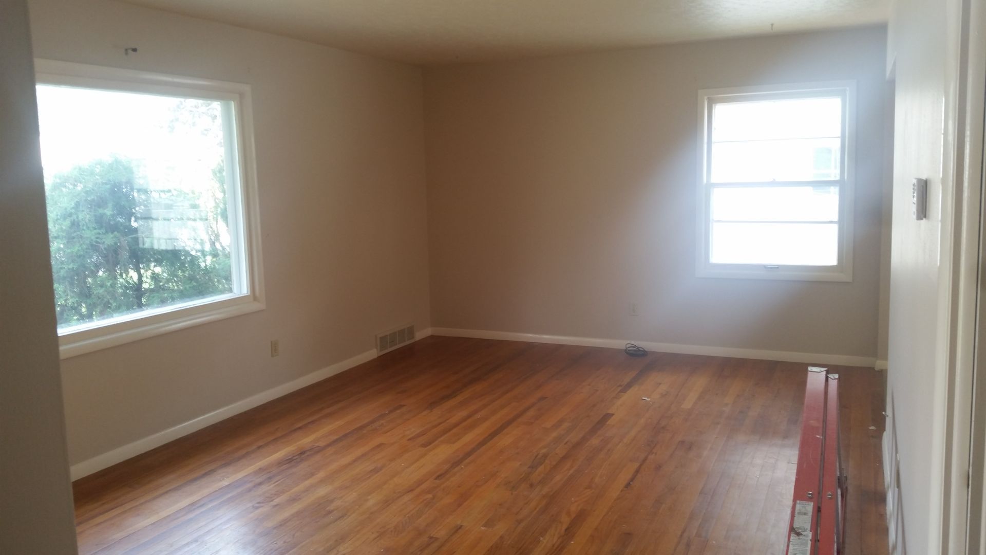 Empty room with hardwood floors, beige walls, and two windows letting in daylight.