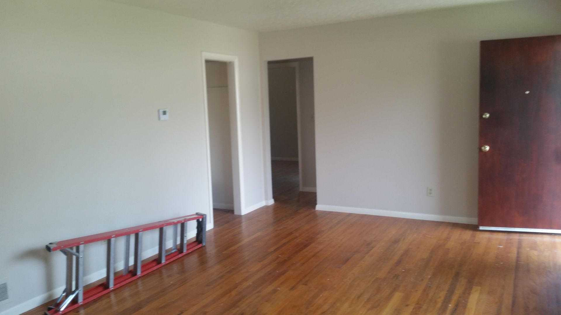 Empty living room with hardwood floors, beige walls, an open doorway, a ladder, and a dark wooden cabinet.
