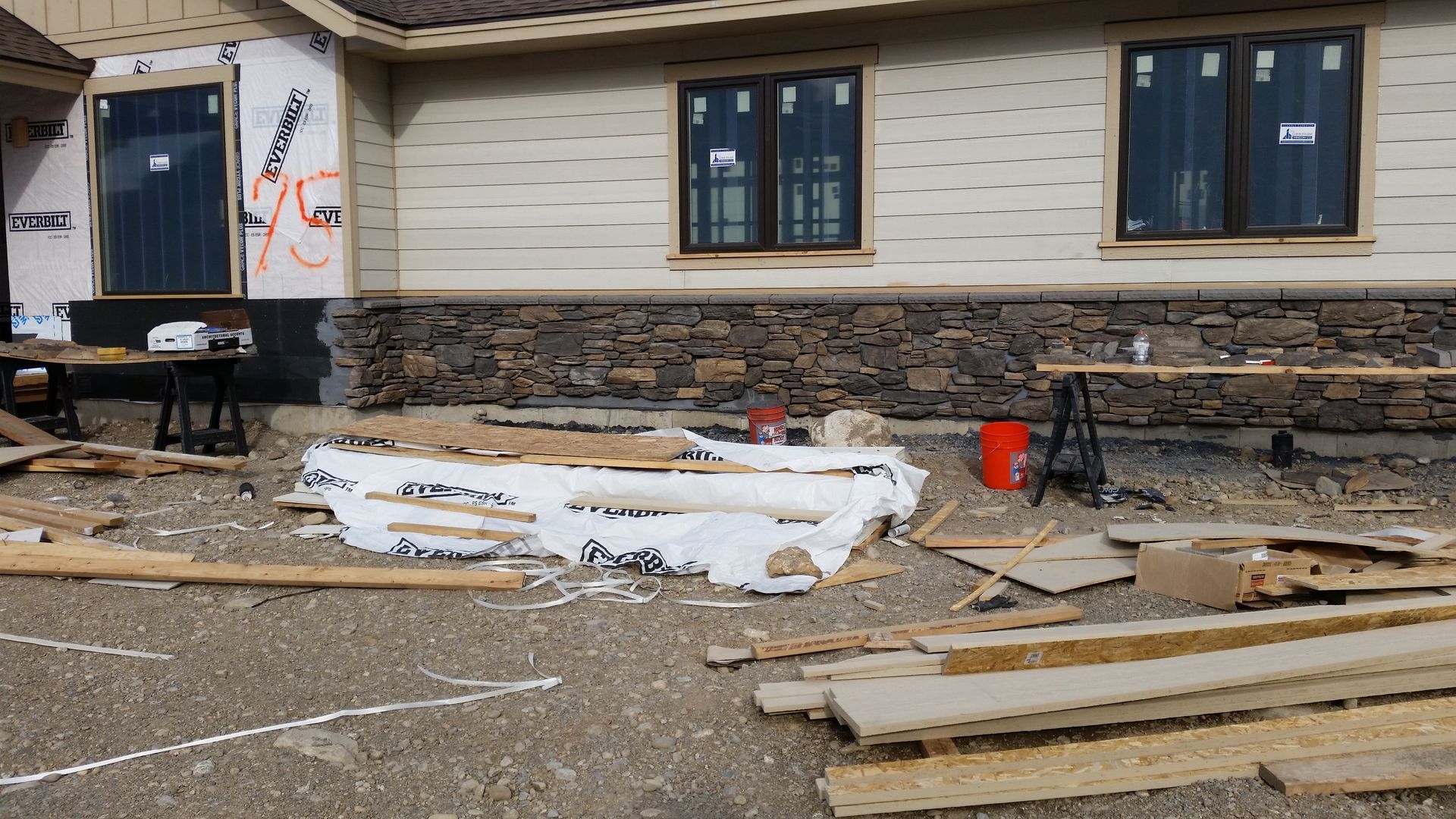 Construction materials and debris scattered outside a beige building with stone foundation and windows