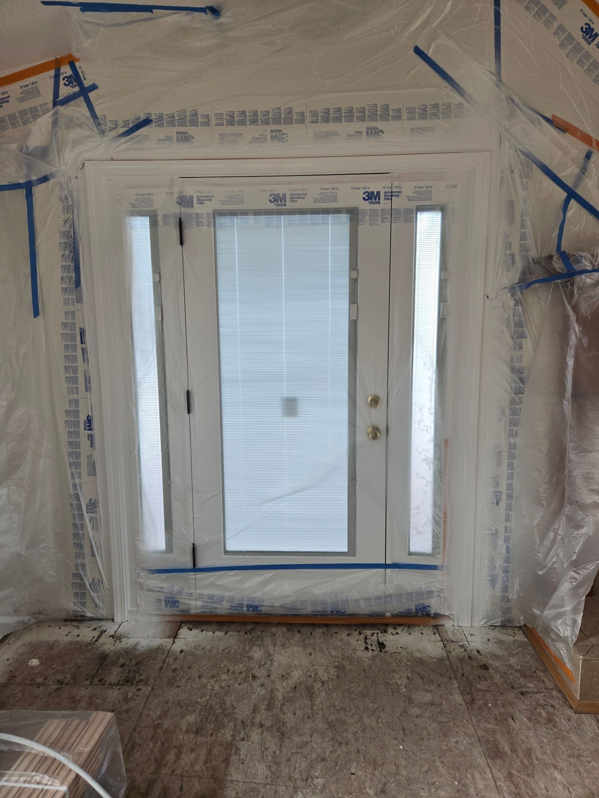 Construction-wrapped patio doors with white frame and blinds, seen in an unfinished room.