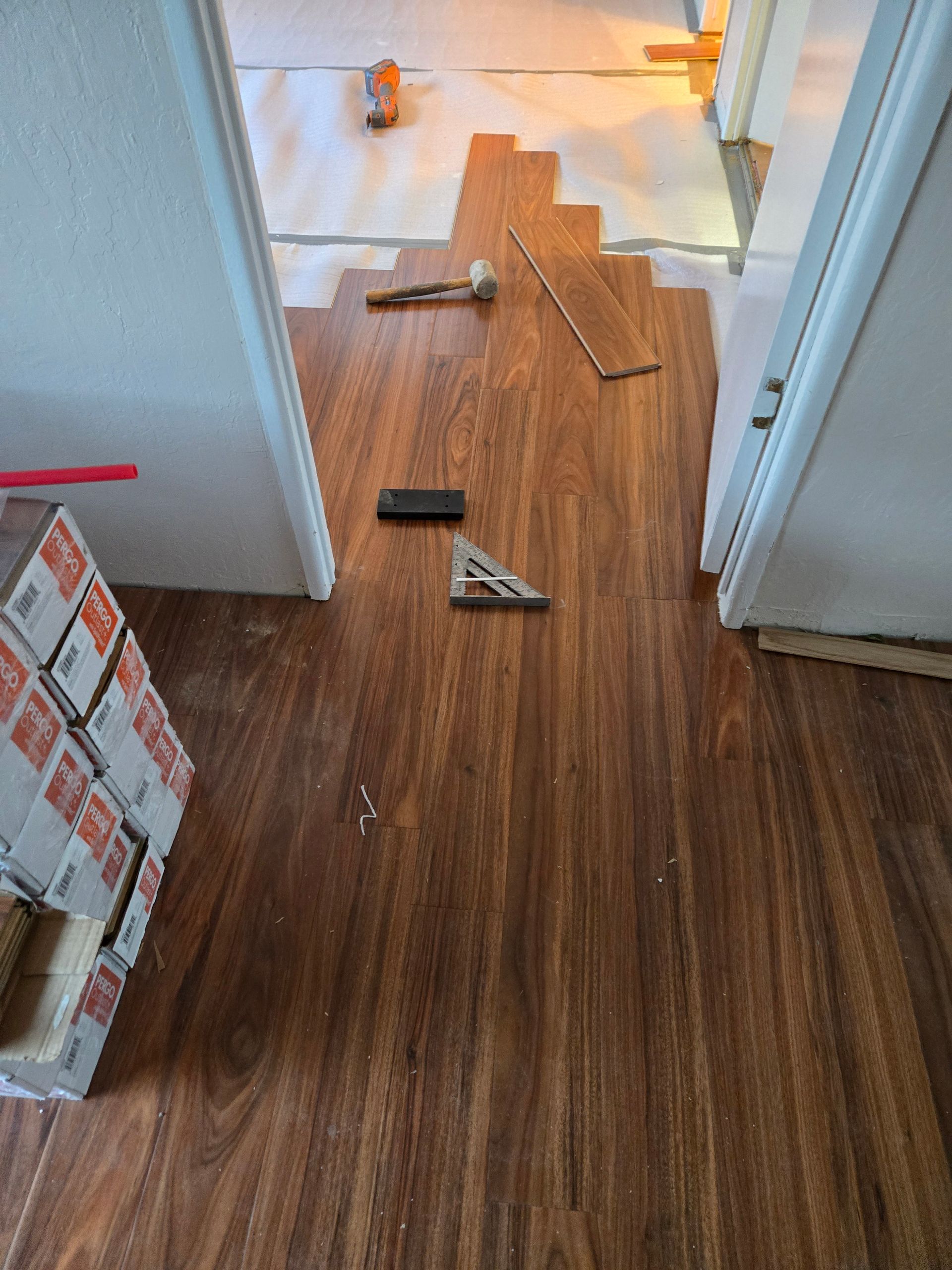 Hallway with damaged hardwood flooring and scattered tools, leading to a bright room ahead.