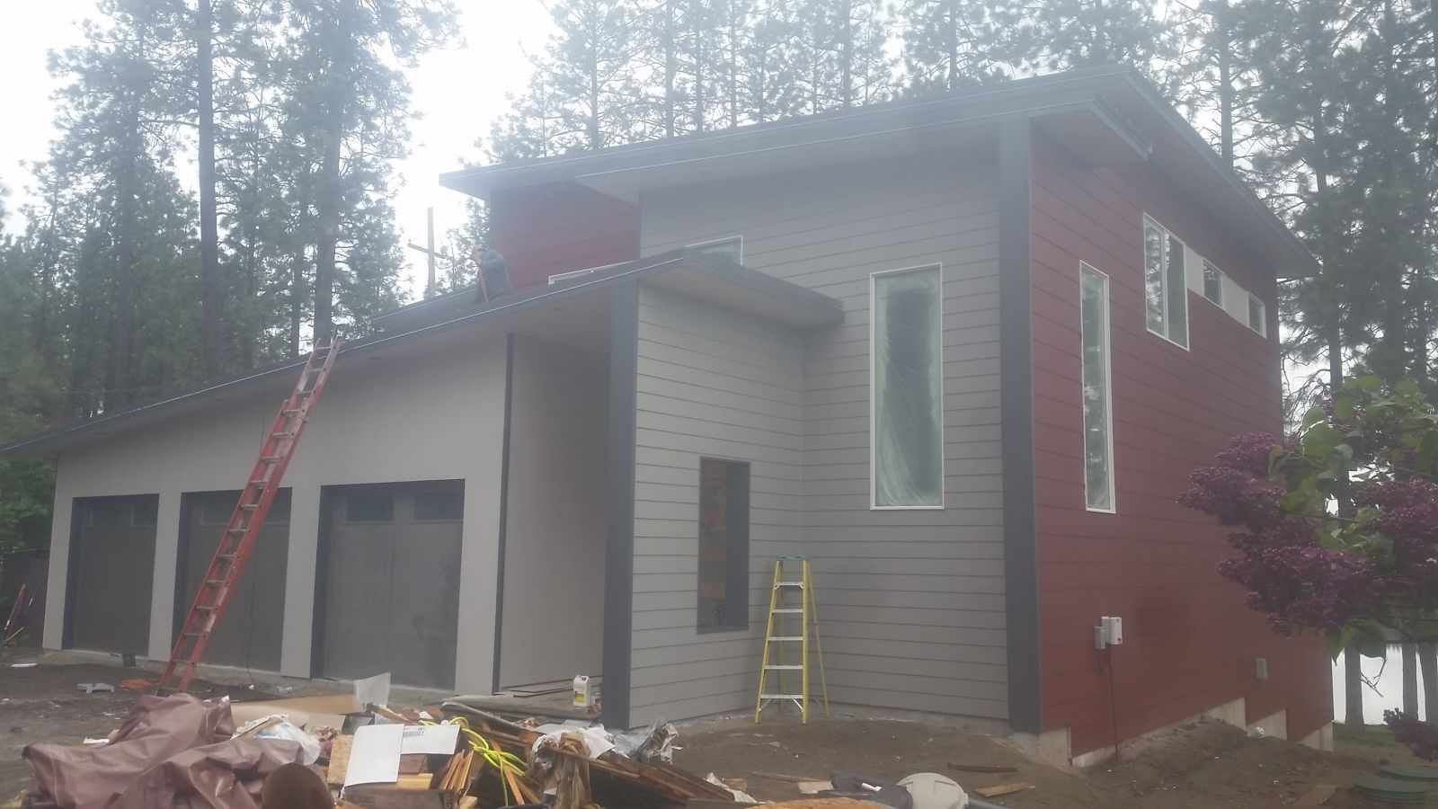 Modern house exterior under construction with red and gray siding, ladders, and trees in the background