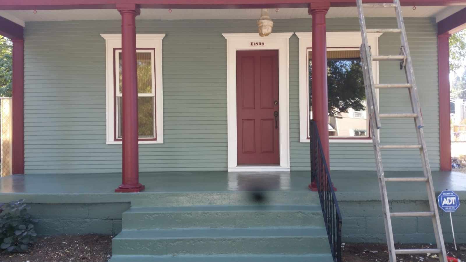 Front porch of a house with green siding, red door, steps, and a ladder leaning on the right side.