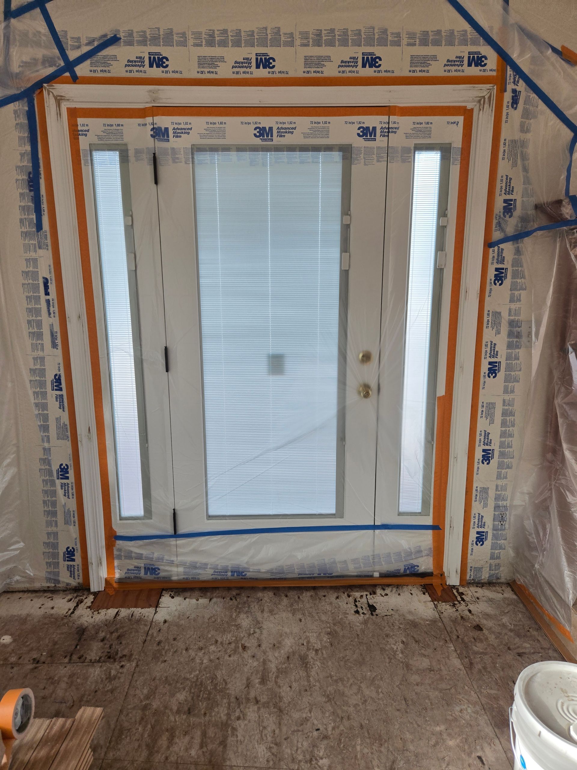Interior under construction with a glass door and sidelights framed by orange tape and plastic sheeting.