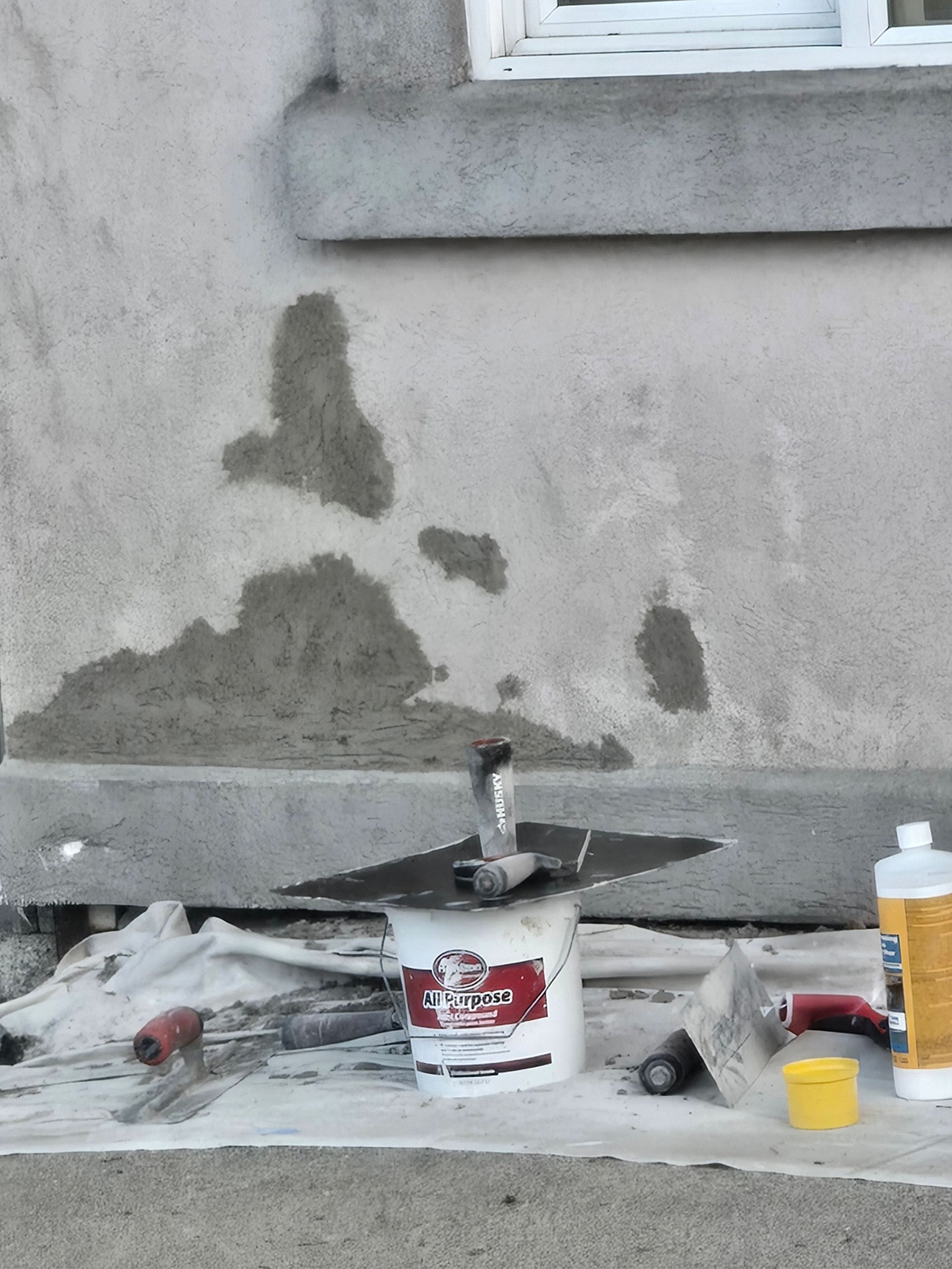 Paint supplies and tools on plastic sheeting below a wall with patchy gray repair spots