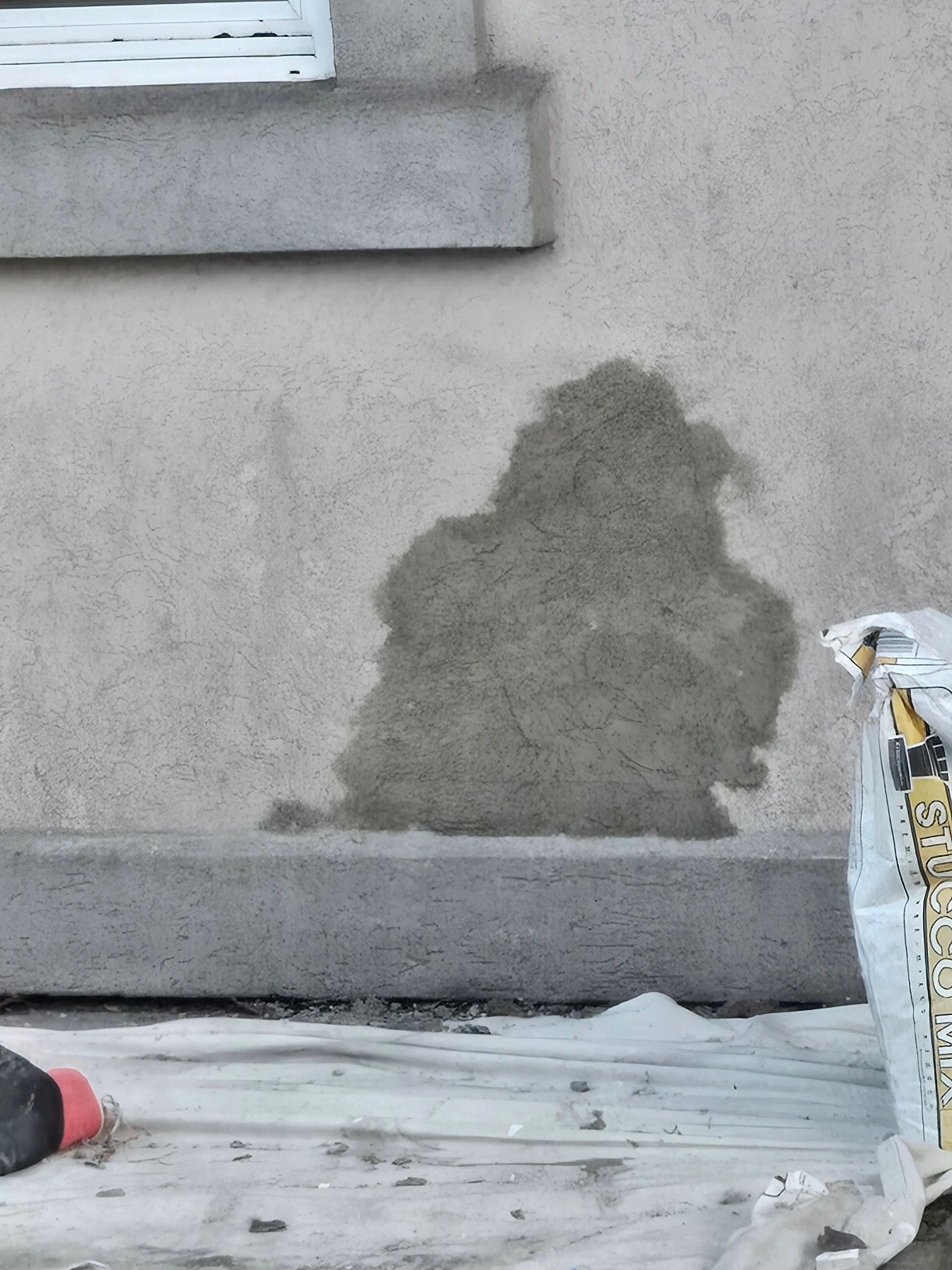 Gray wall with a damp stain near the base, beside a window and construction debris on the ground