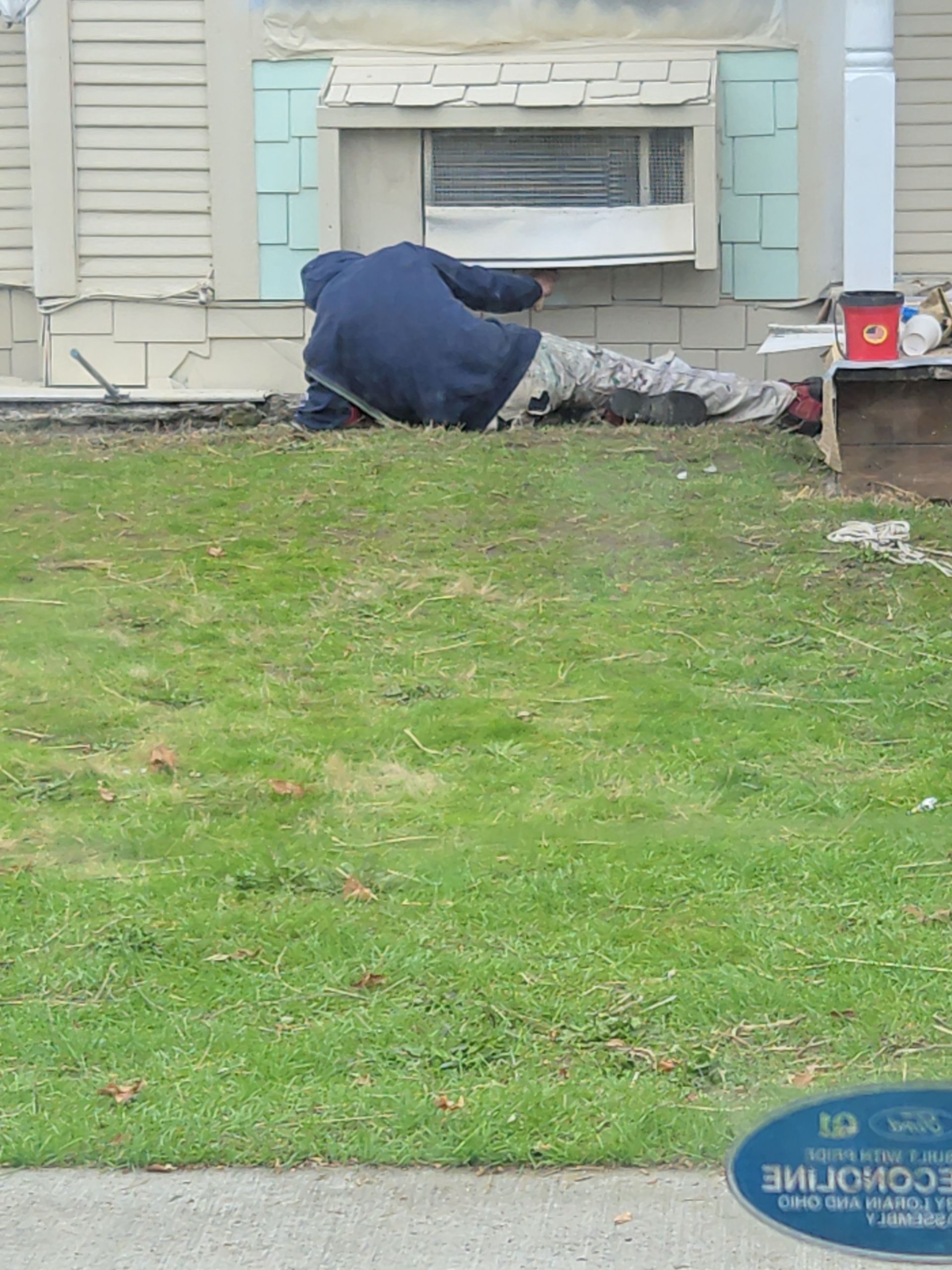 Person lying on the ground beside a house wall under a basement window, inspecting something near the foundation.