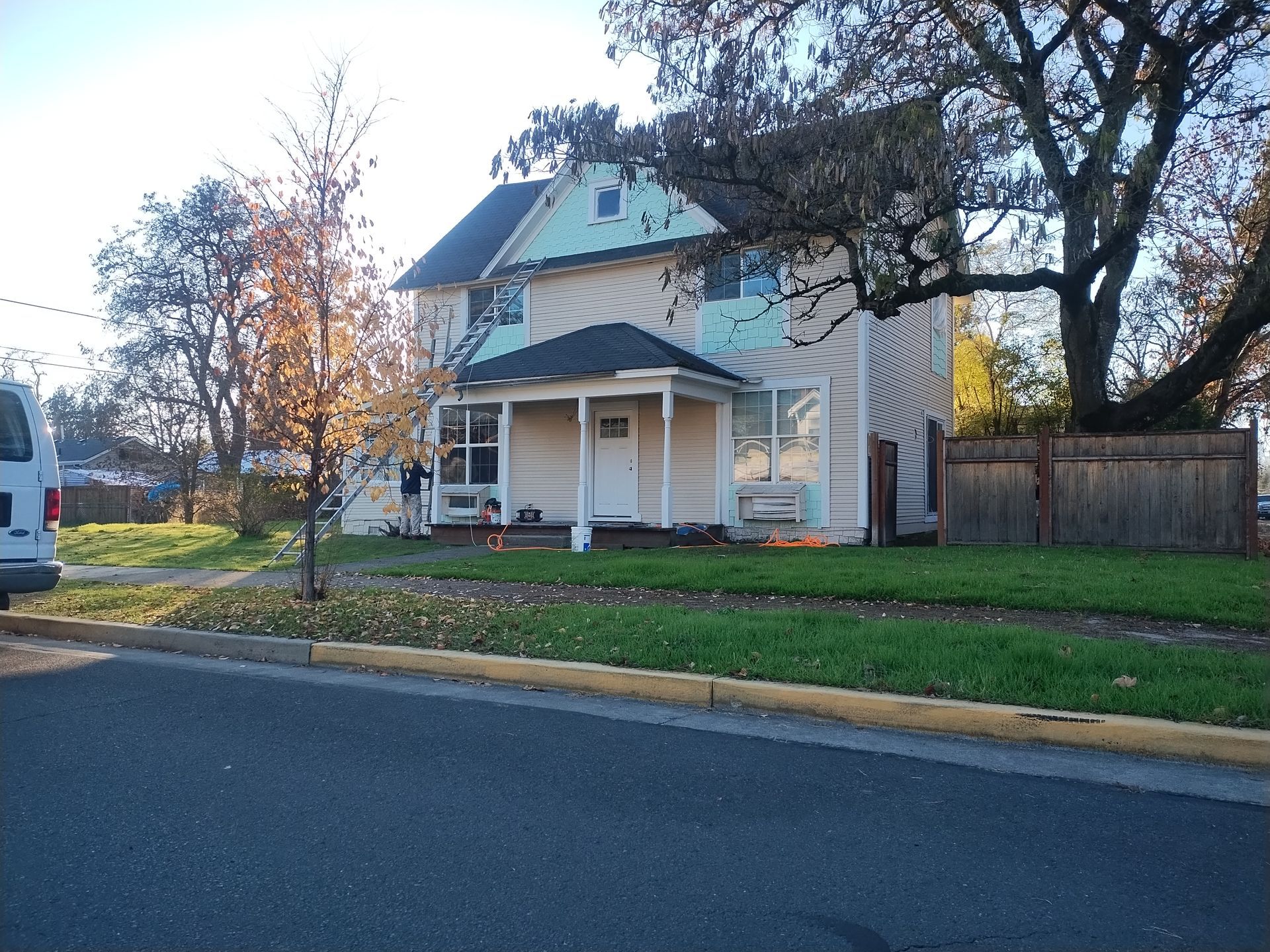 Two-story beige house with a porch and blue roof beside a quiet street in autumn.