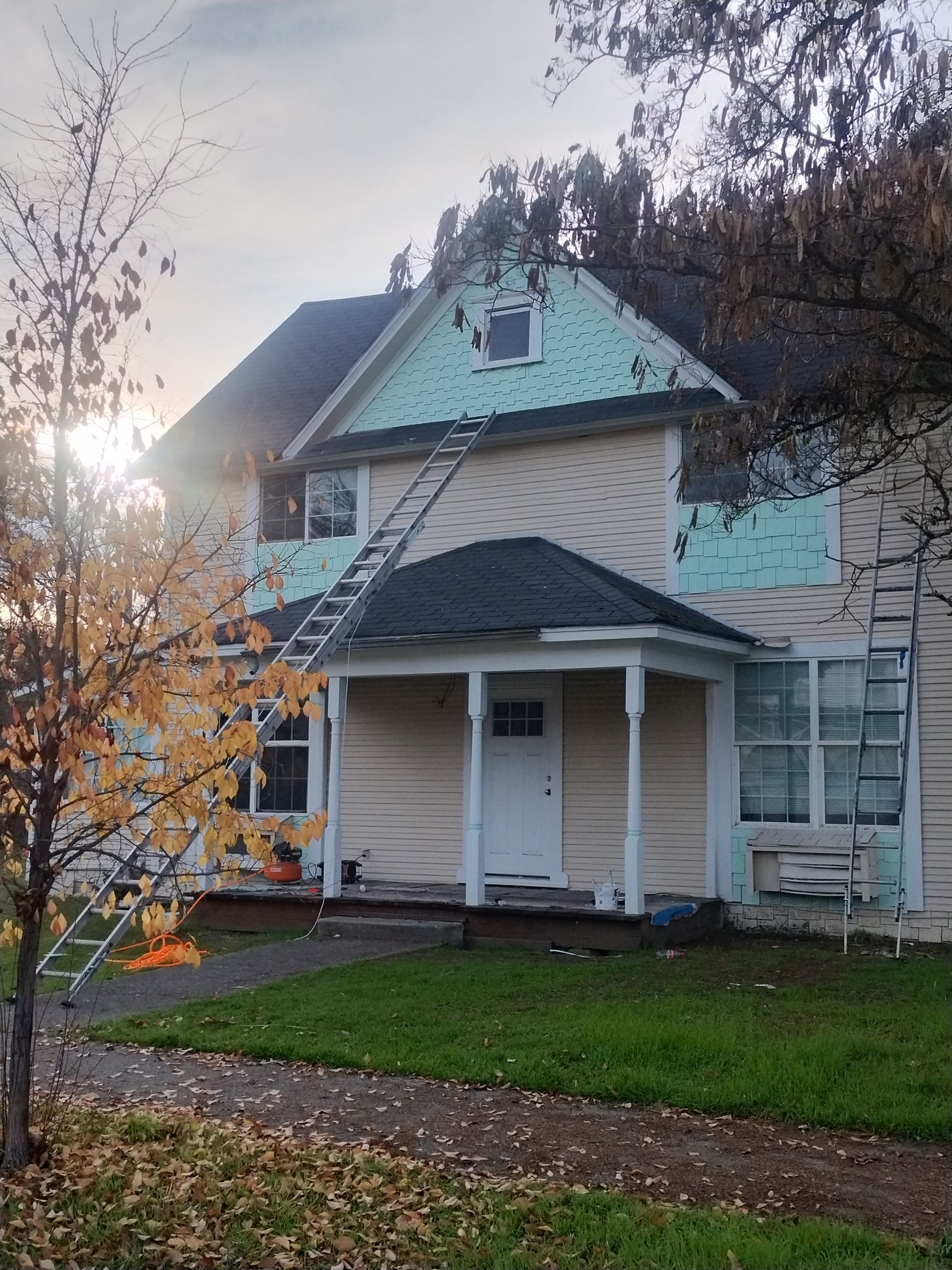 Two-story white house with teal roof and porch, autumn leaves, and sunlight at the left edge.