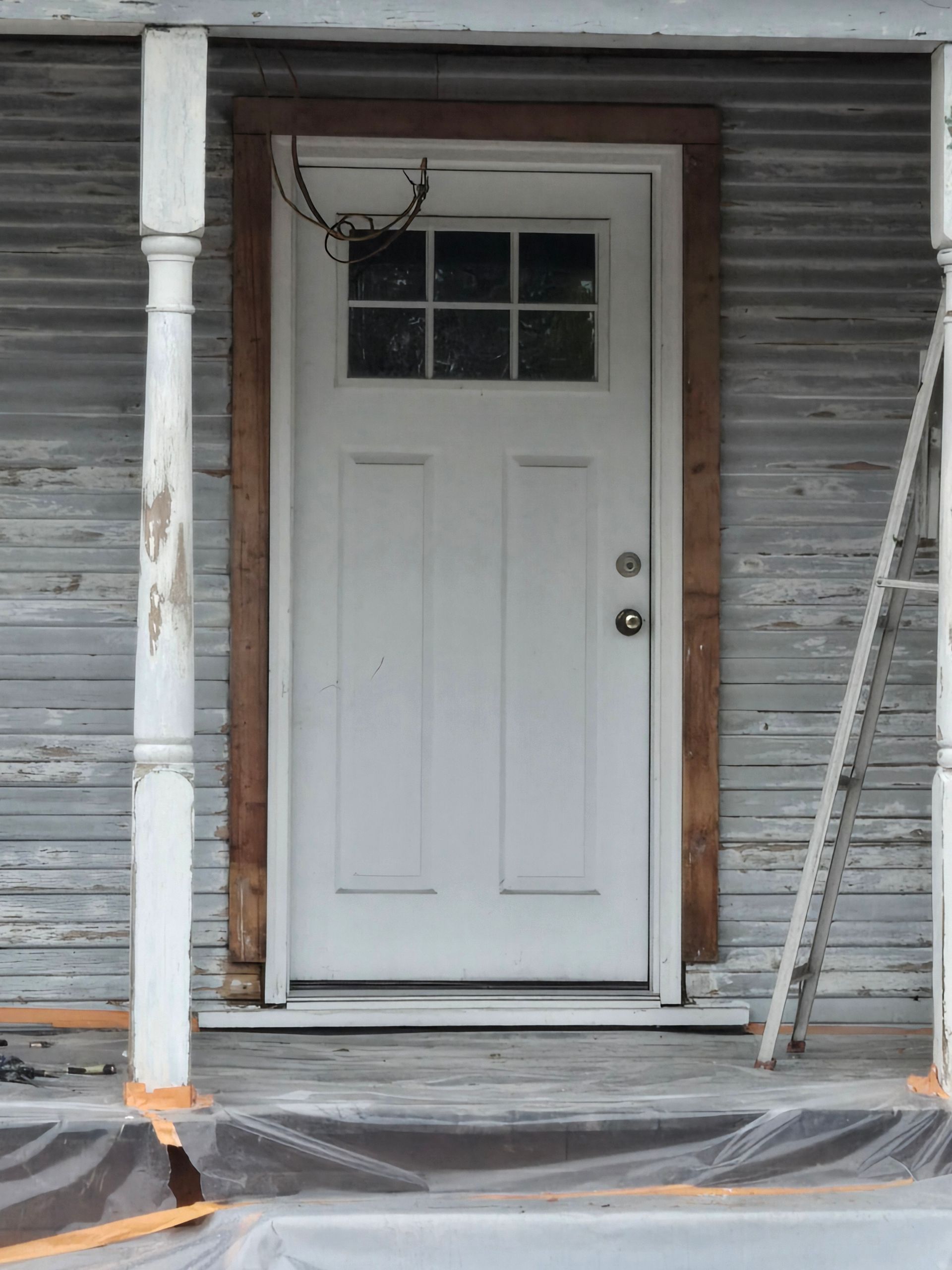 White front door in a weathered siding exterior, with a porch under renovation and plastic sheeting on the ground