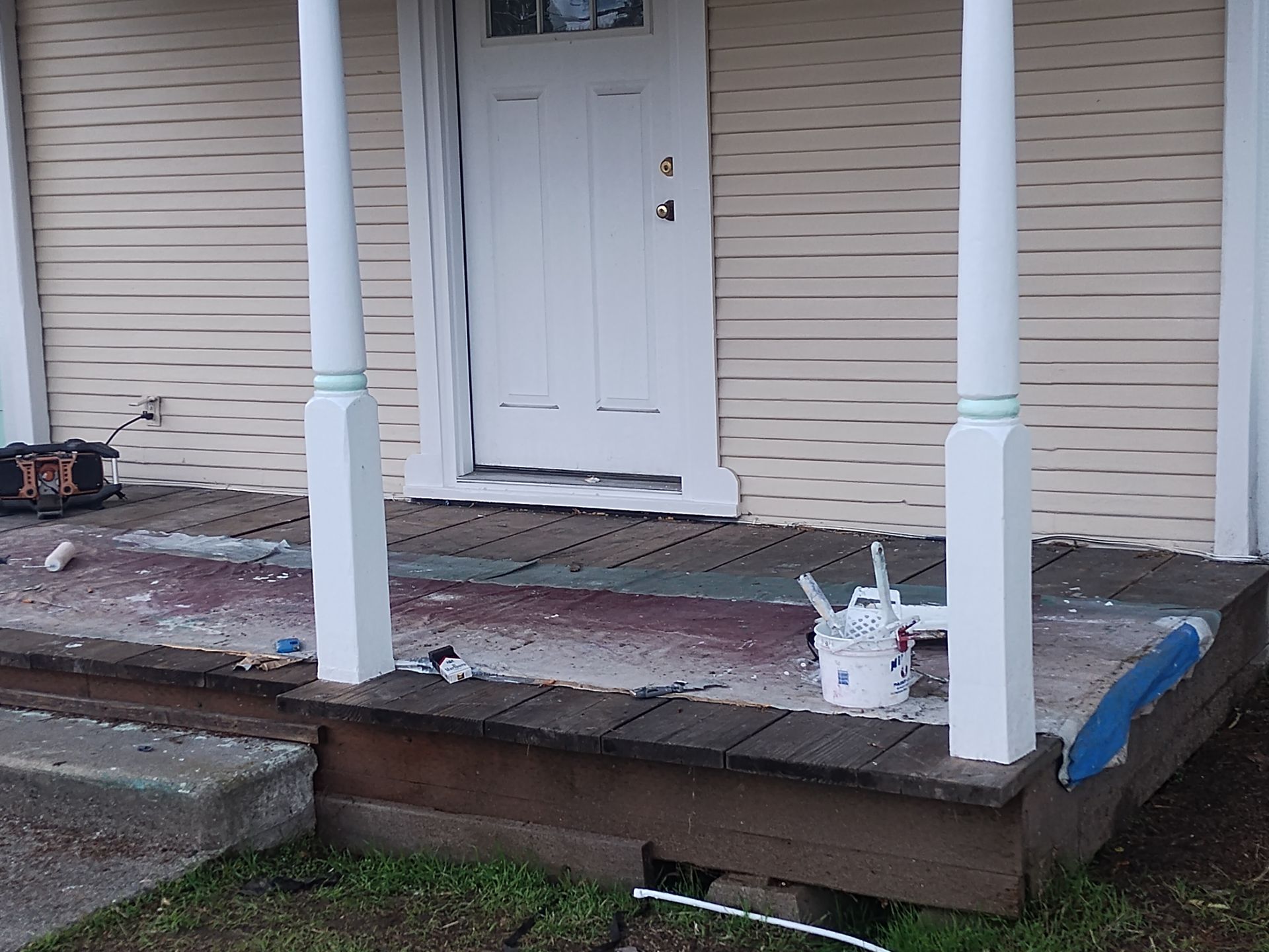 Front porch with white door, columns, and a weathered wooden deck with paint cans.
