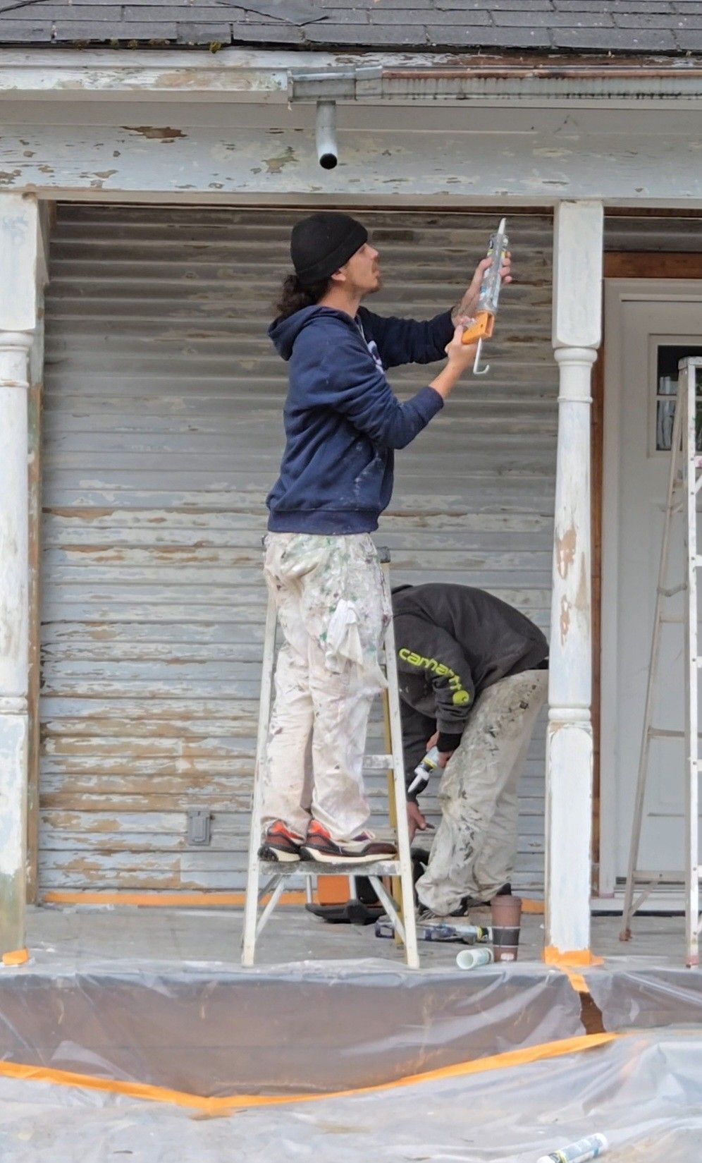 Two workers painting a porch exterior, one on a ladder and one crouched below.