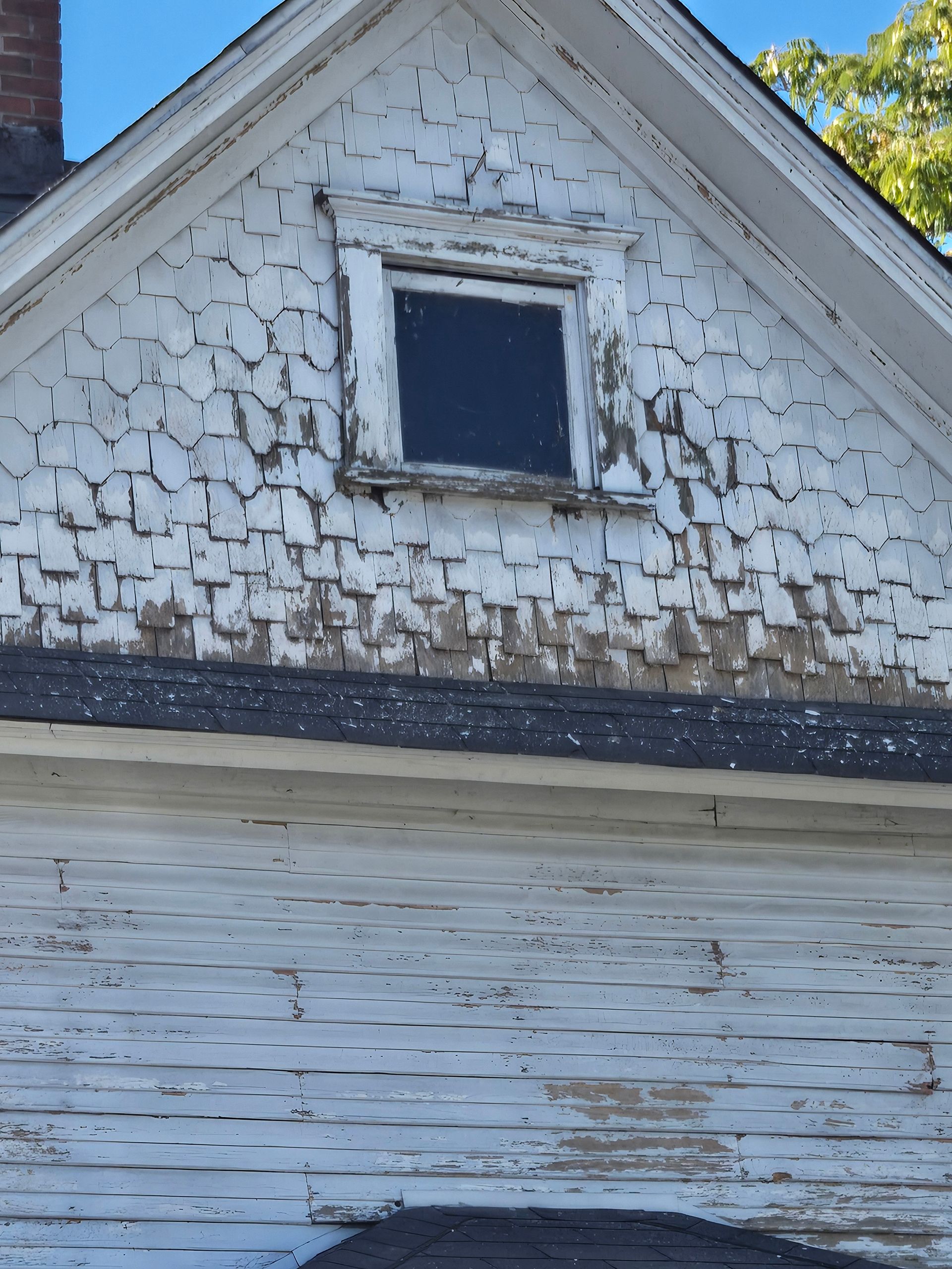Peeling white paint on a house gable with a small dark attic window
