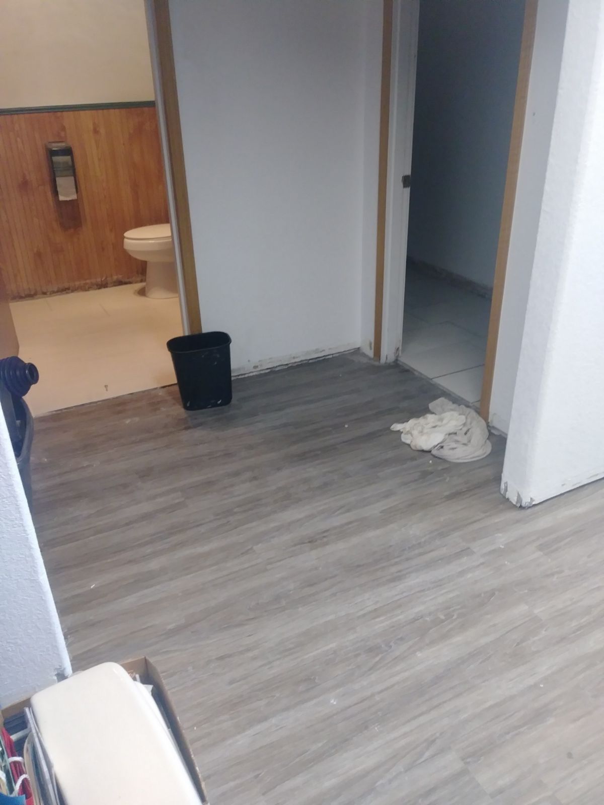 Empty room with gray wood floor, white walls, open doorway, and a black trash bin near the wall