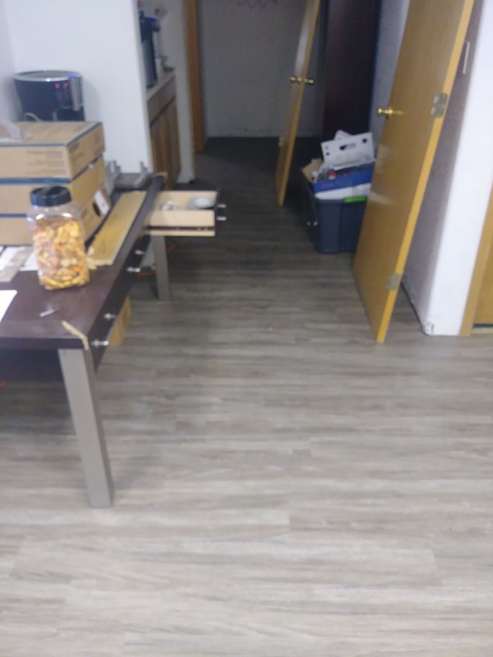 Small office room with tables, boxes, and an open door, viewed down a hallway with wood flooring.