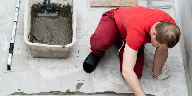 Person in a red shirt kneeling on a rooftop, spreading concrete beside a bucket of wet cement.