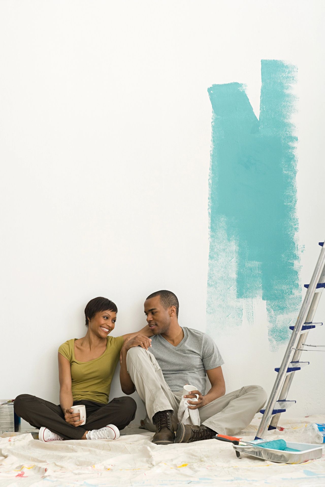Two people sitting on the floor painting a wall teal, with a ladder nearby.