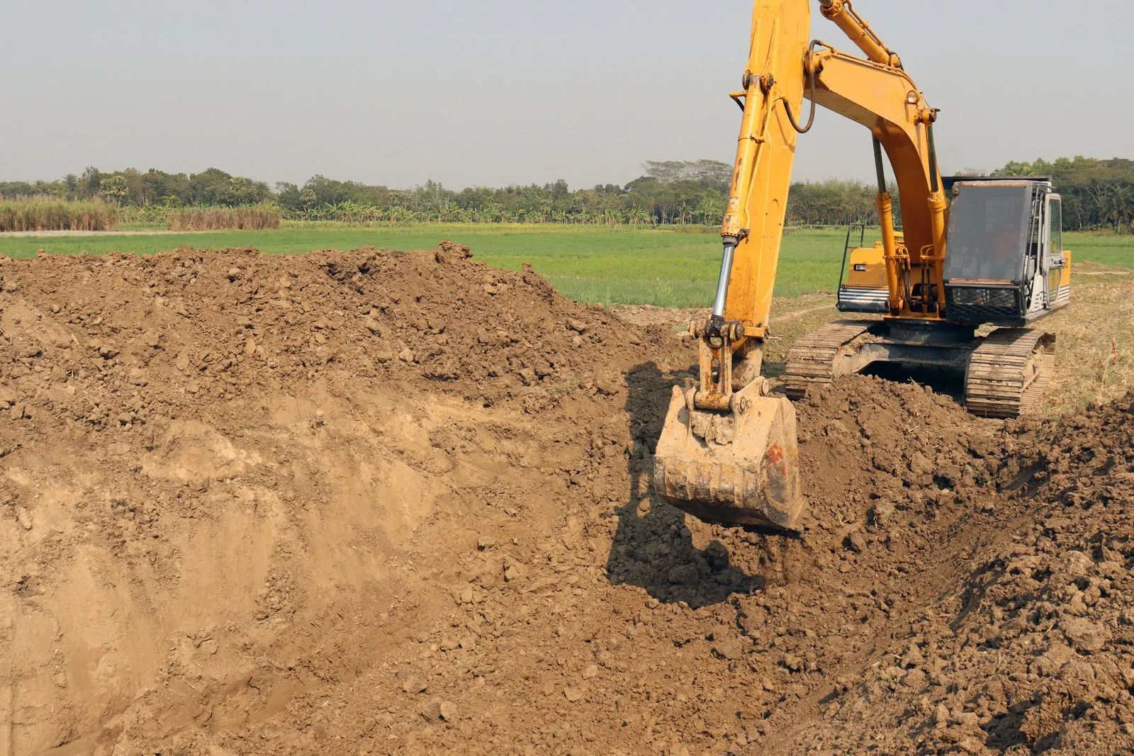 Yellow excavator digging a dirt trench in a rural field under a clear sky