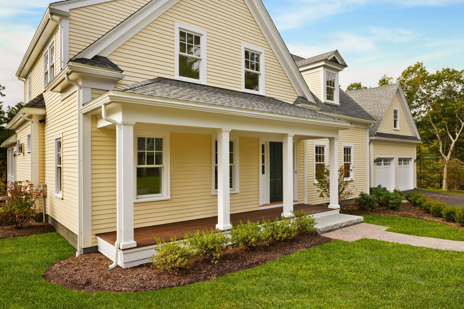 Yellow house with white trim, front porch, and two-car garage; green lawn, blue sky.