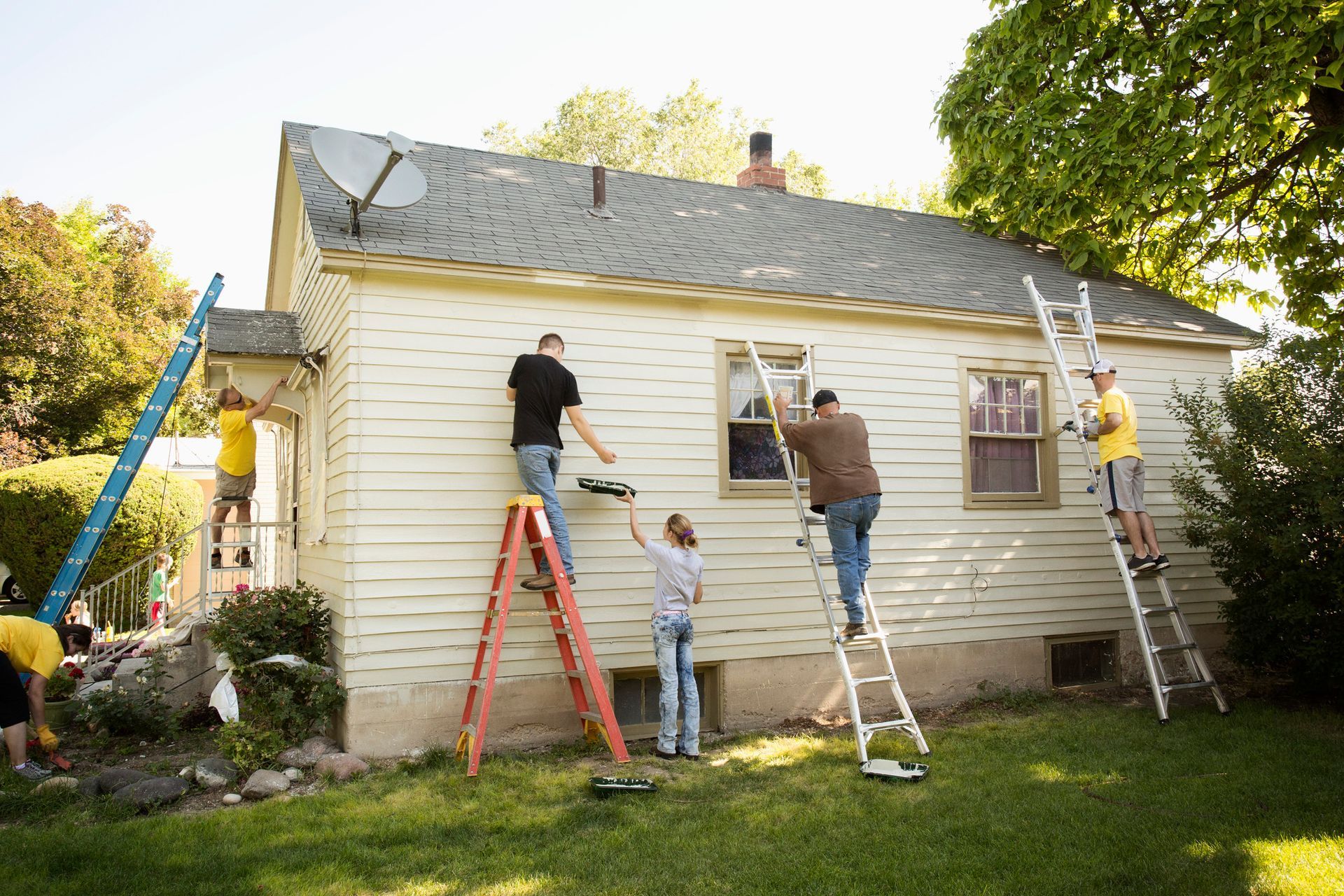 People on ladders painting the exterior of a white house with green grass and trees in the yard.