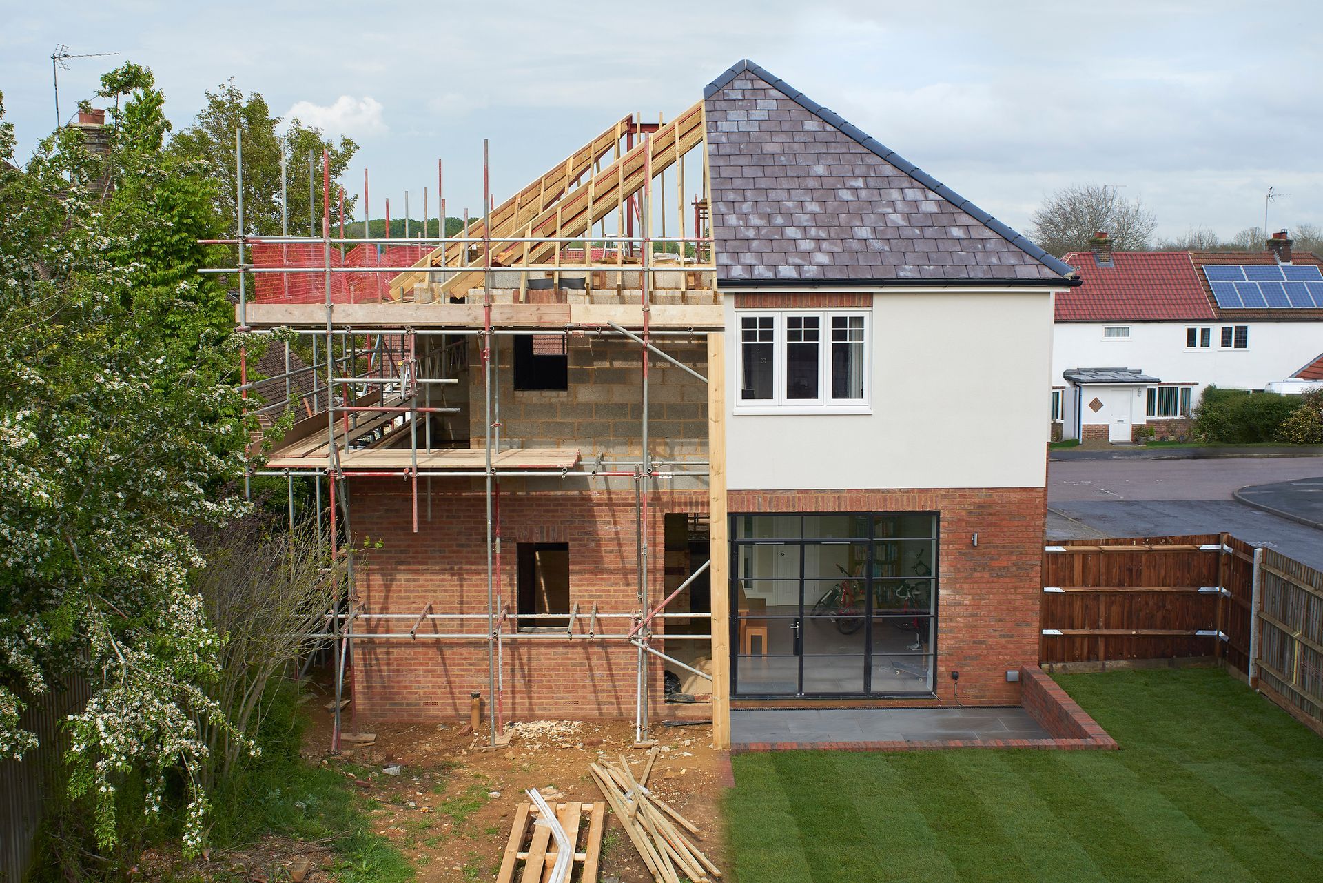 House under construction, half finished, with scaffolding, brick and wood, adjacent to a finished section with gray tile and glass doors.