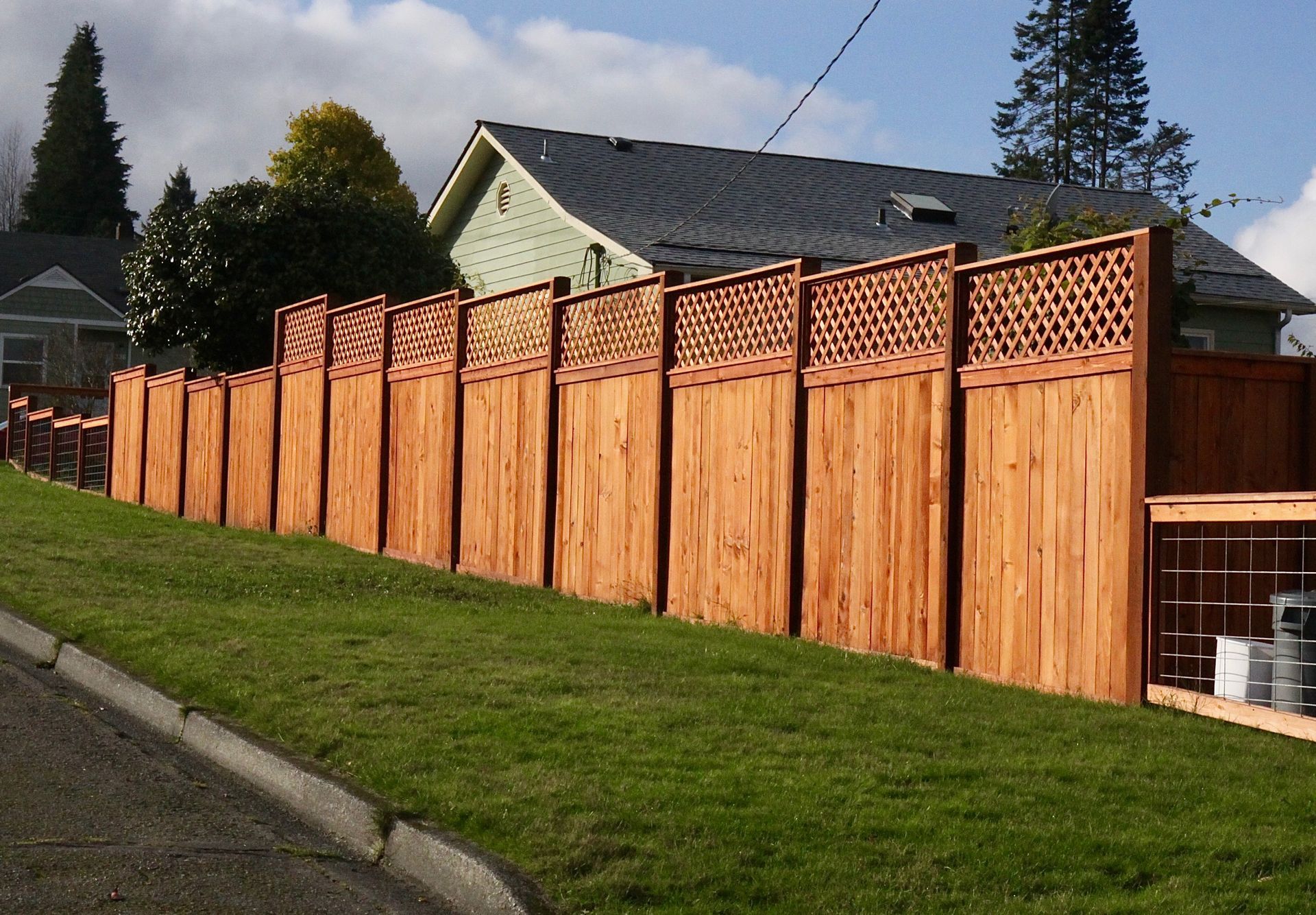 Wooden fence in front of a green lawn, separating the yard from a street. Lattice tops, sunny.