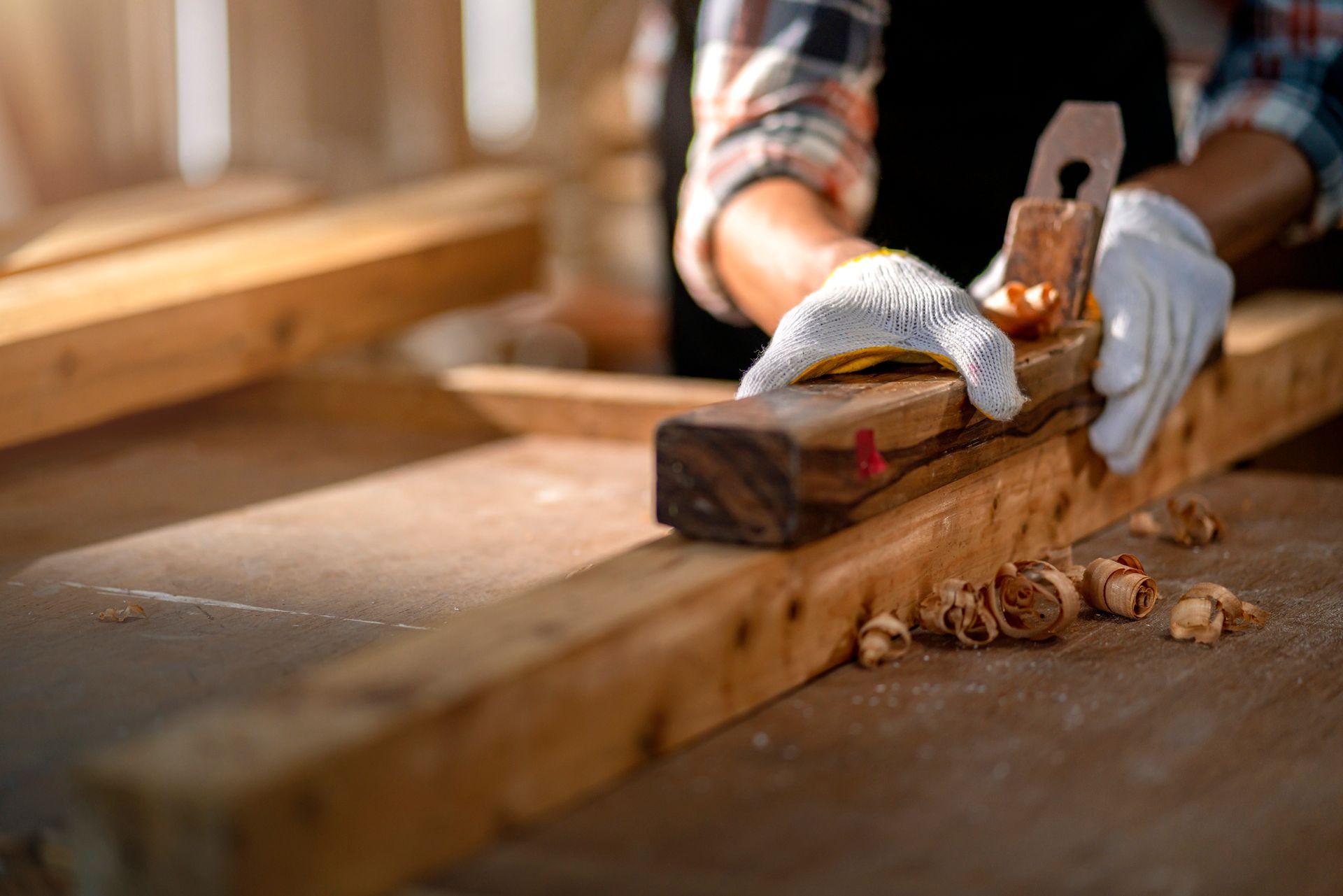 Carpenter planing a wooden plank, wearing gloves, in a workshop.