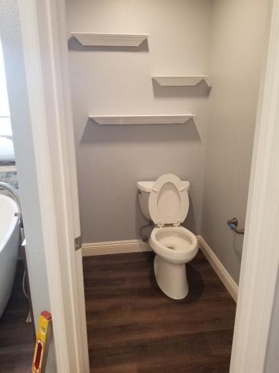 Toilet in a small, light gray room with brown flooring, and white shelves.